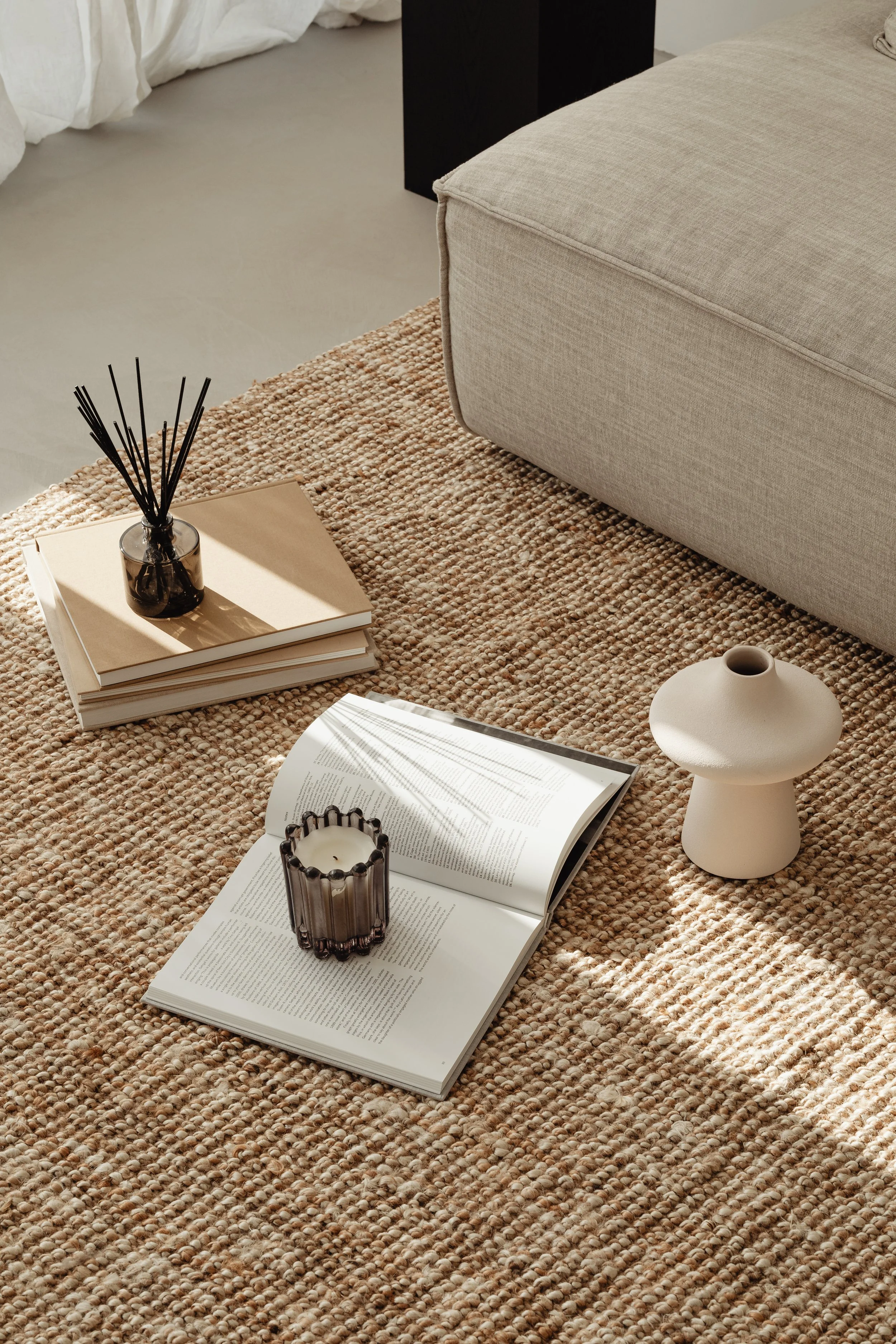 A cozy setting on a textured beige rug with an open book, a glass candle holder, a small black and white candle, a closed book with a black reed diffuser, a beige ceramic vase, a beige sofa, and a black side table in the background.