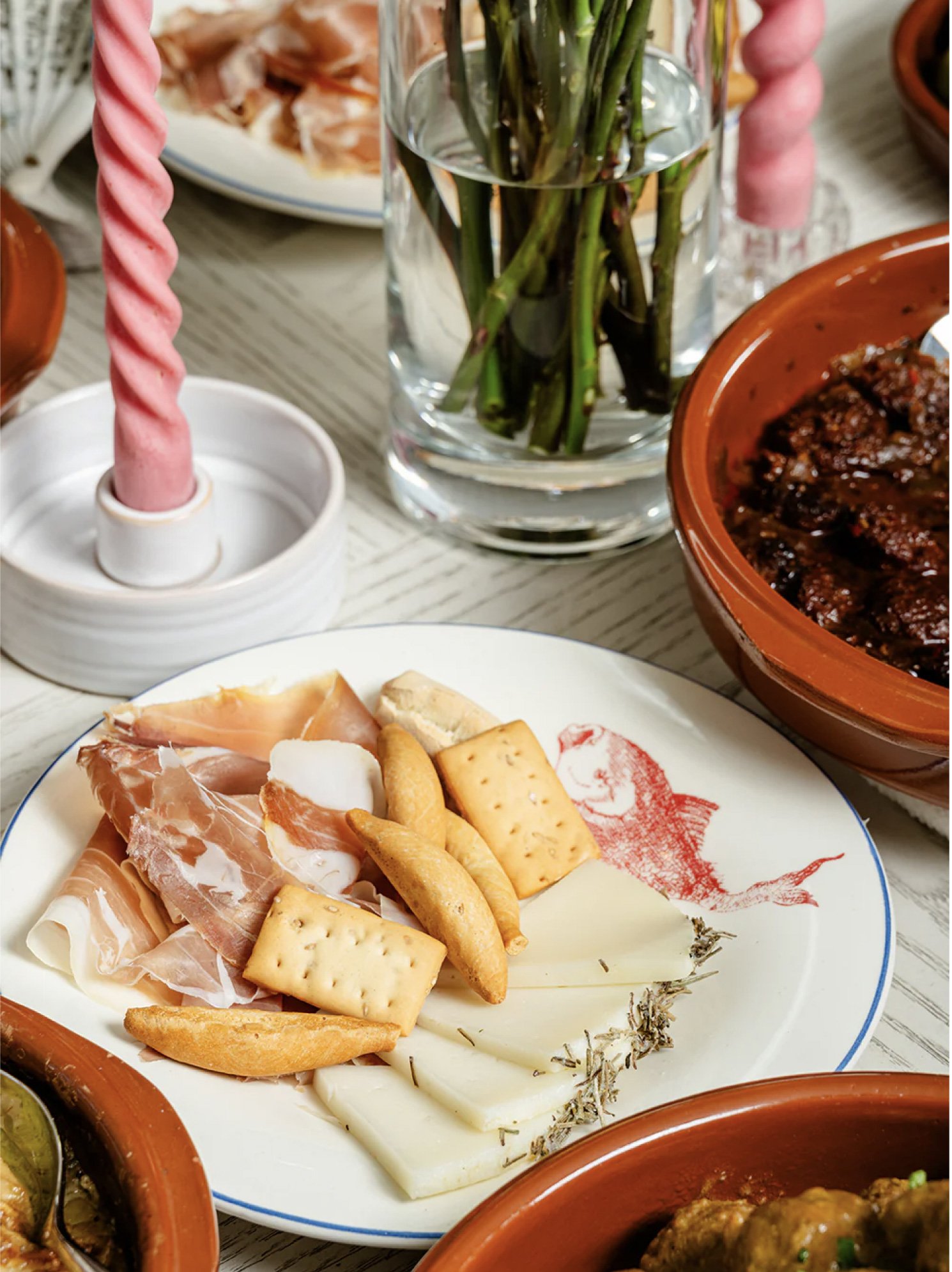 A table with various Mexican dishes, including a plate of sliced cured ham, cheese, crackers, and bread. A small dish of guacamole can be seen in the lower left corner, and a glass vase with green plants is in the background.
