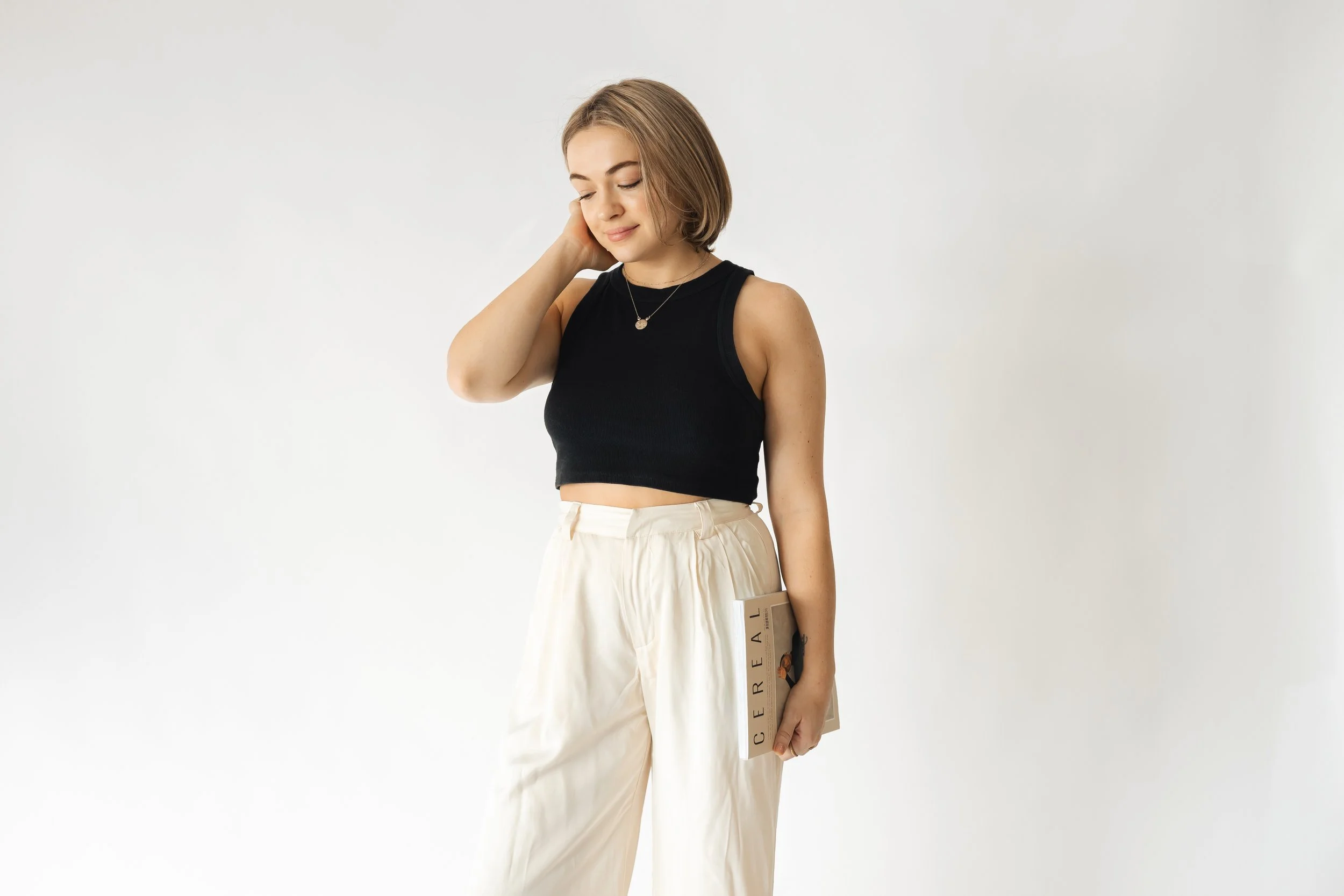 A young woman with shoulder-length blonde hair, wearing a black sleeveless crop top and beige high-waisted pants, standing against a plain white background, holding a magazine in her left hand and gently touching her face with her right hand, with her eyes closed.