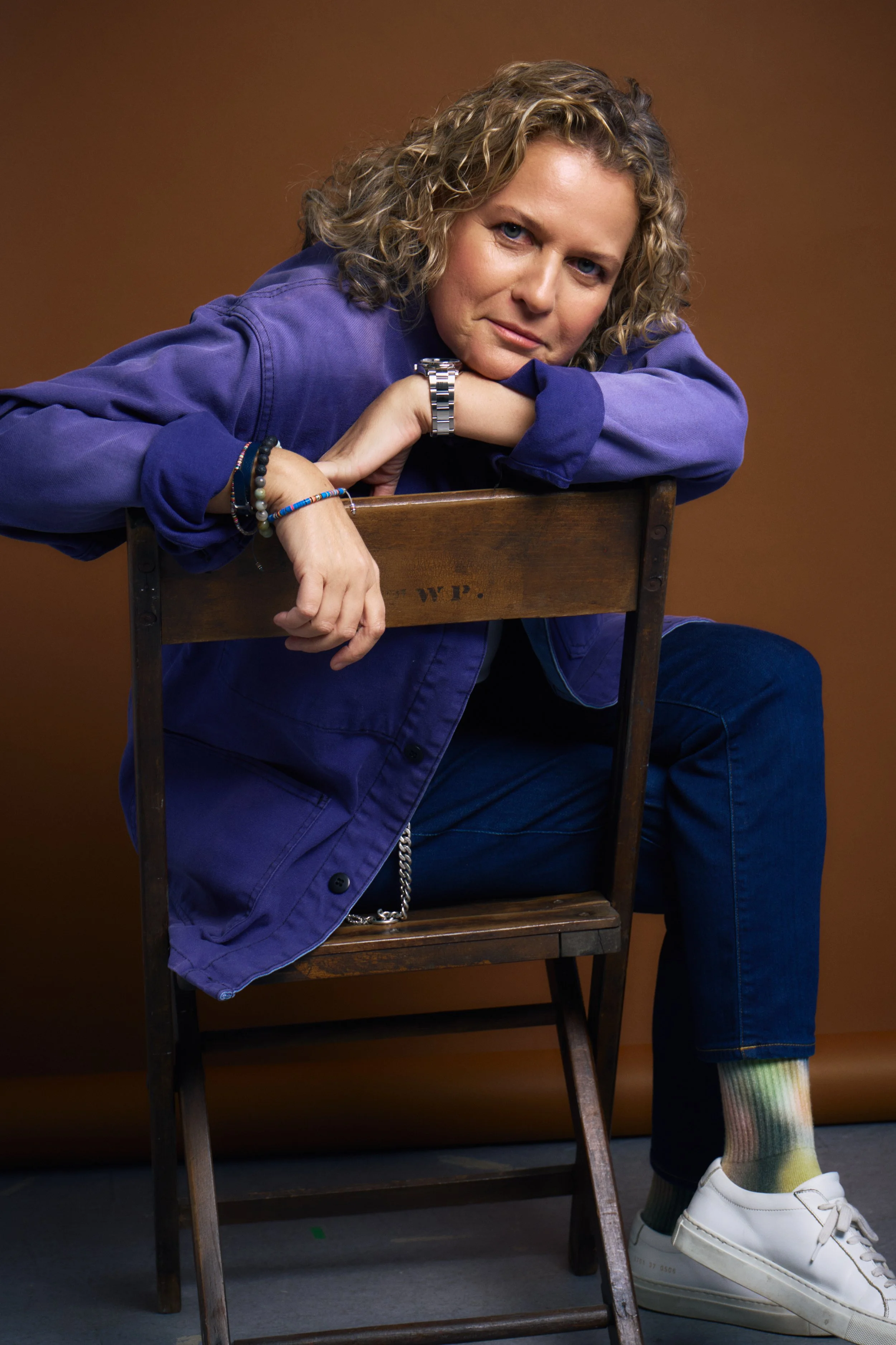 A woman with curly blonde hair wearing a purple jacket, sitting on a wooden chair, resting her arms on the back of the chair, with her gaze directed at the camera. She is wearing a watch, bracelets, and multicolored socks with white sneakers. The background is plain brown.