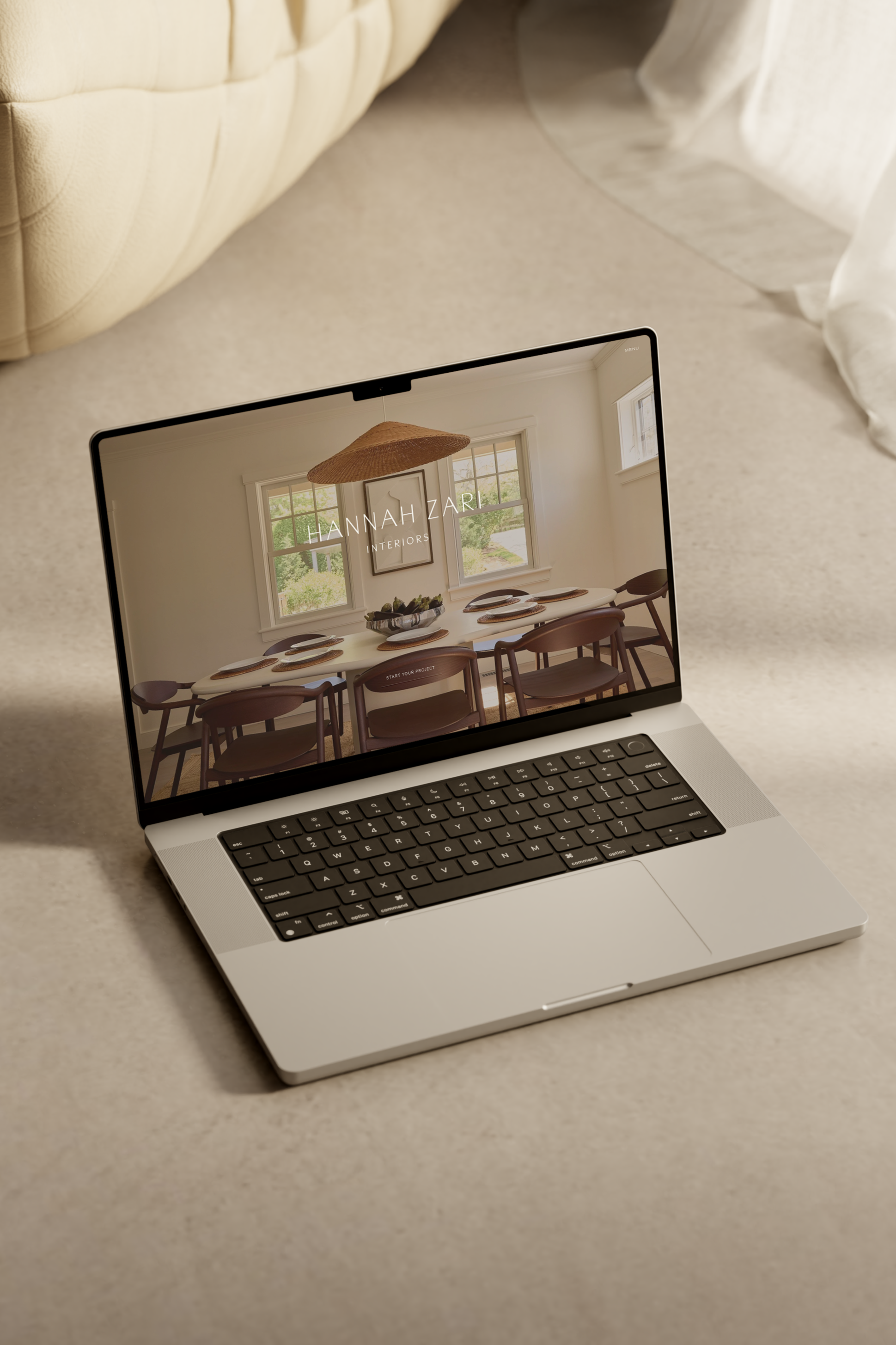 A laptop resting on a beige carpet, displaying a webpage for Hannah Zari Interiors with a dining room image.