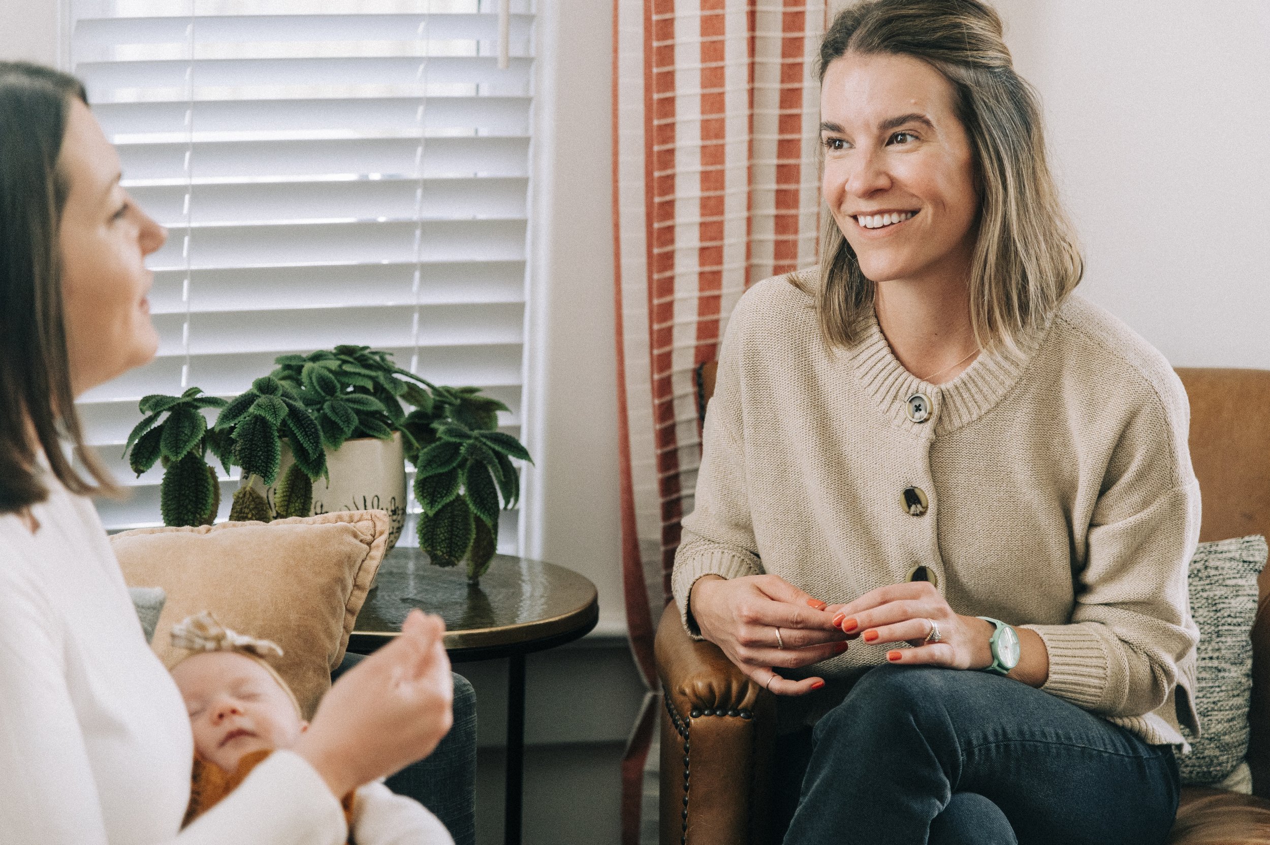 Two women sitting and smiling, one holding a sleeping baby, in a cozy home with window blinds, a potted plant, and cushions.