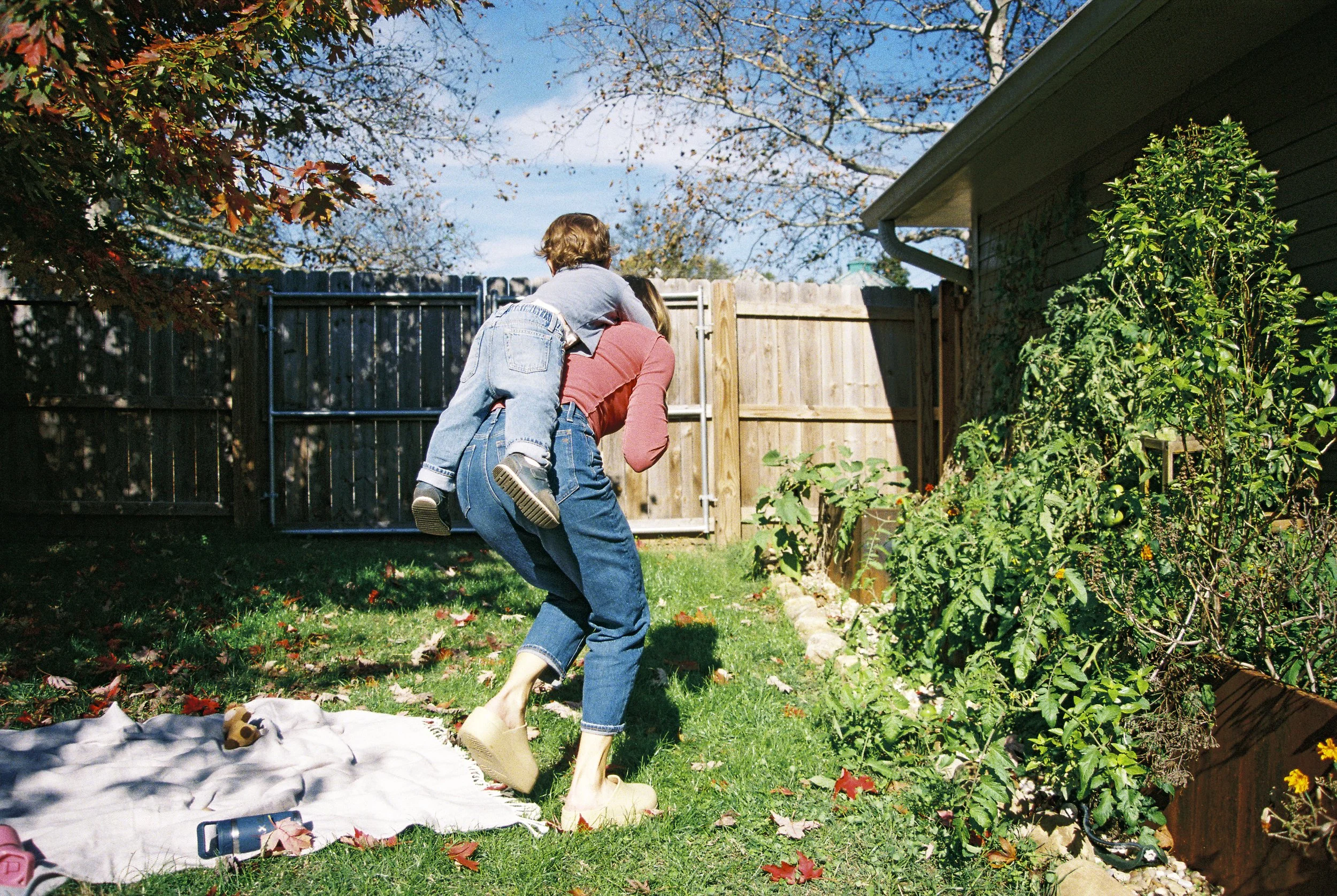A woman outside in a backyard carrying a young child on her back, as they walk on grass near a garden with plants and trees, with a wooden fence and gate in the background on a sunny day.