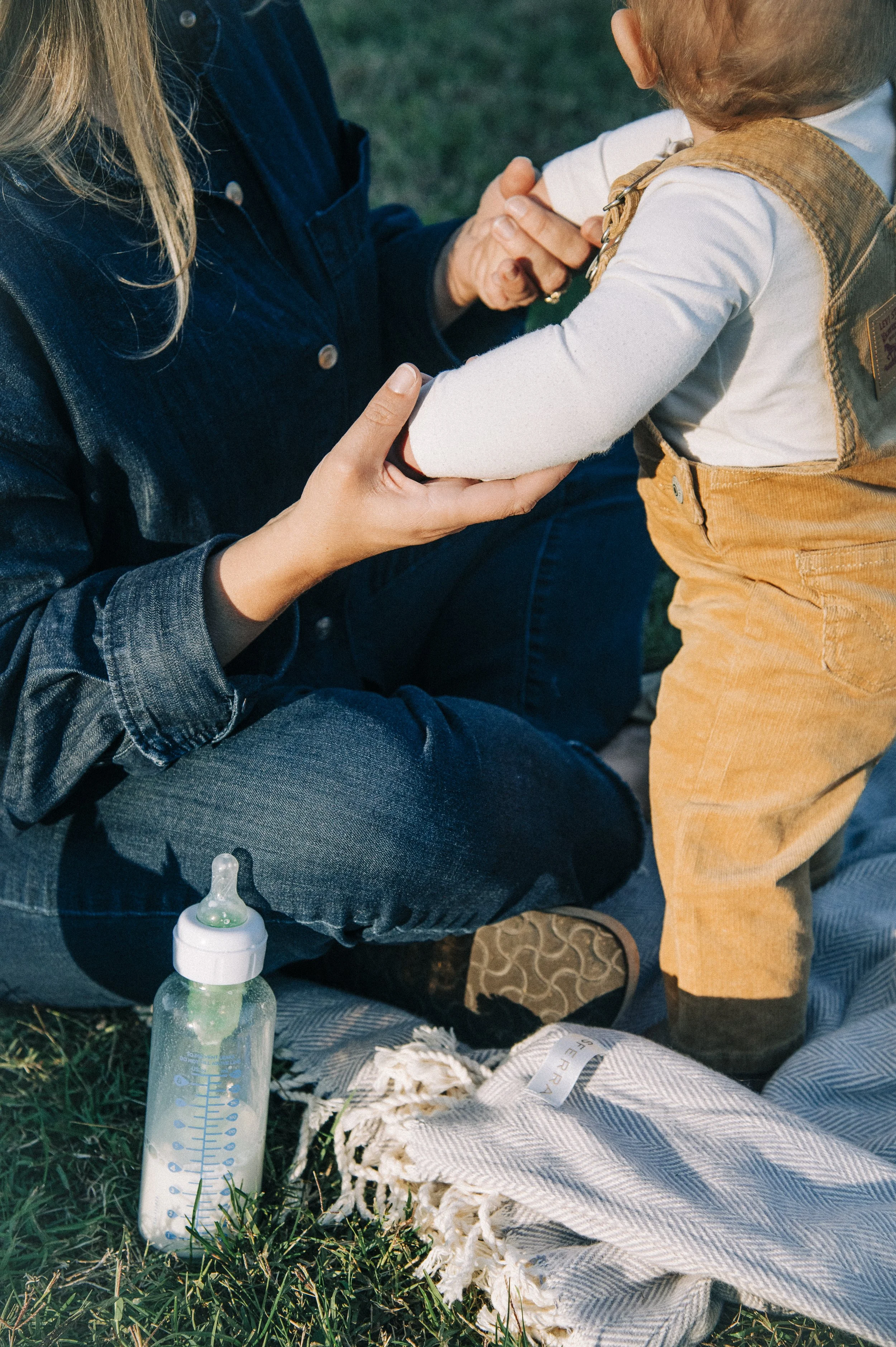 A woman sitting on the grass holding a child's arm, preparing to give a bottle, with a baby bottle and a cloth on the grass nearby.