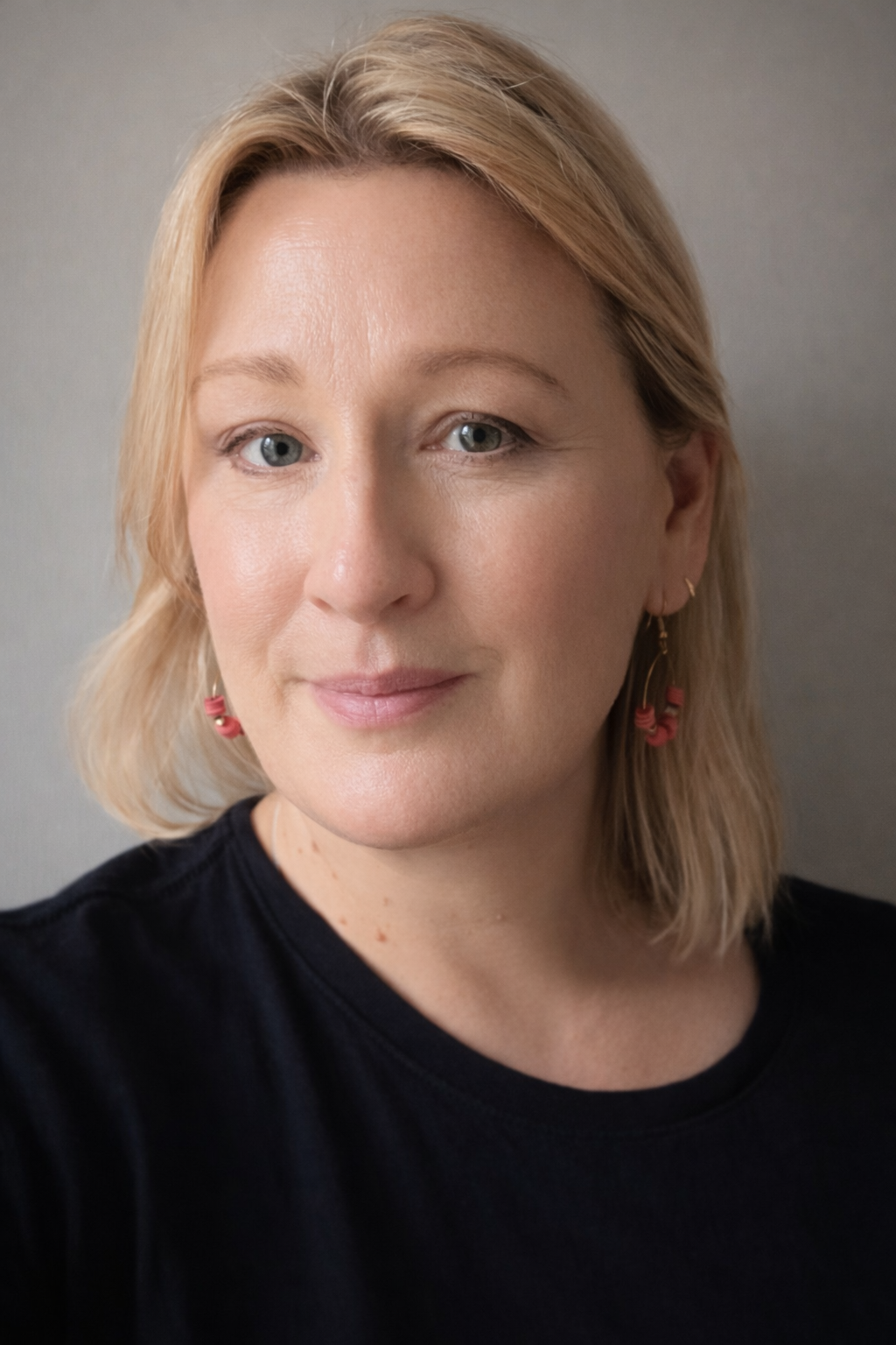A woman with blonde hair, blue eyes, and light skin looking at the camera. She is wearing a black top and pink earrings with small red beads.