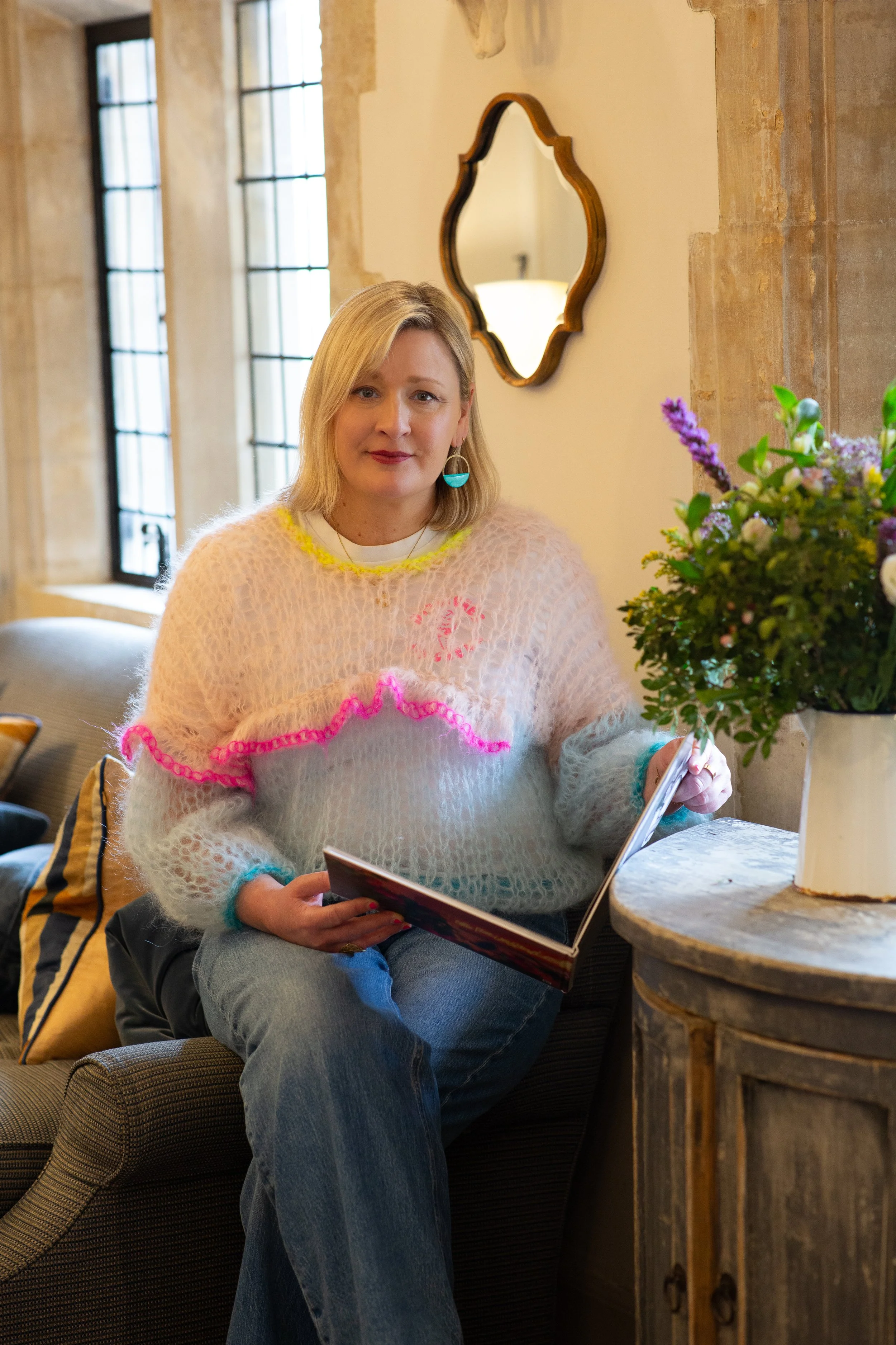 A woman sitting on a brown sofa with a patterned cushion, holding a book, wearing a colorful fuzzy sweater, blue jeans, and teardrop earrings, in a cozy room with large windows and a stone wall. There is a large bouquet of purple, yellow, and white flowers on a round wooden side table beside her.