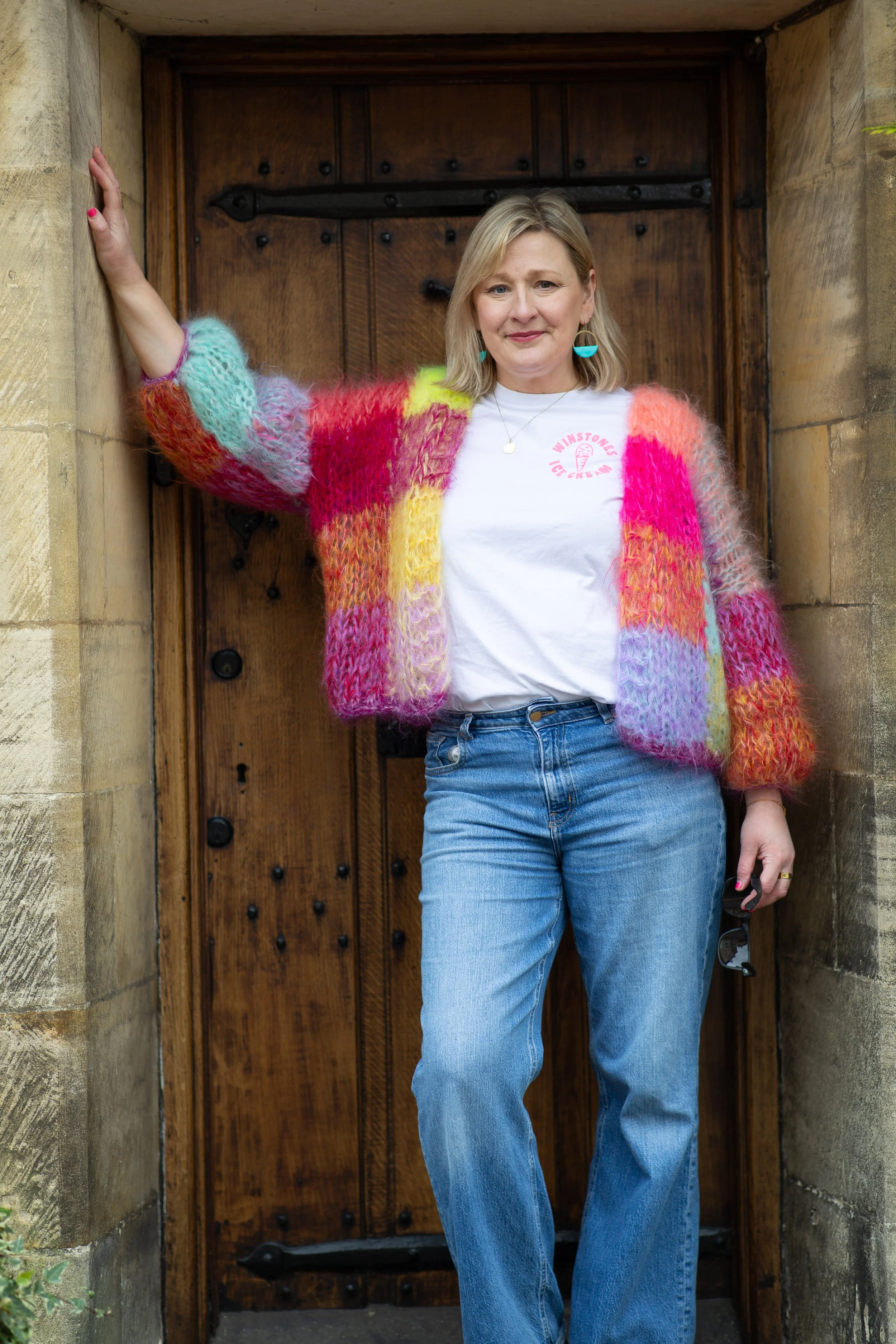 Woman standing outdoors in front of a wooden door, wearing a colorful knitted cardigan, white T-shirt, and blue jeans, holding sunglasses in her right hand.