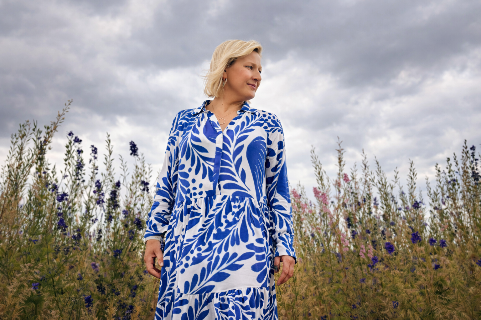 A woman with blonde hair wearing a long blue and white patterned dress standing in a field of purple and pink flowers under a cloudy sky.
