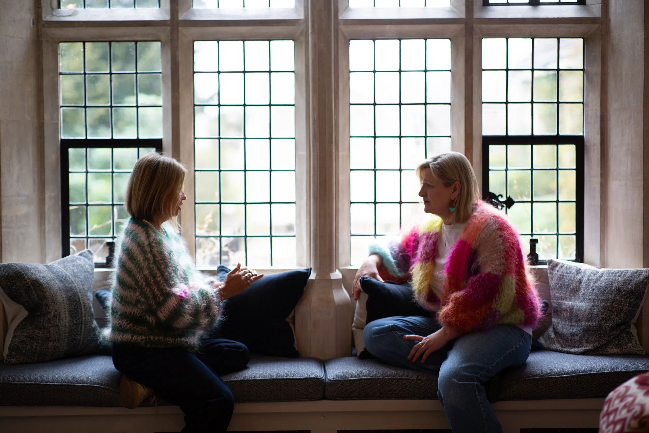 Two women sitting on a window seat, facing each other, having a conversation in a cozy room with large grid-style windows and a stone interior.