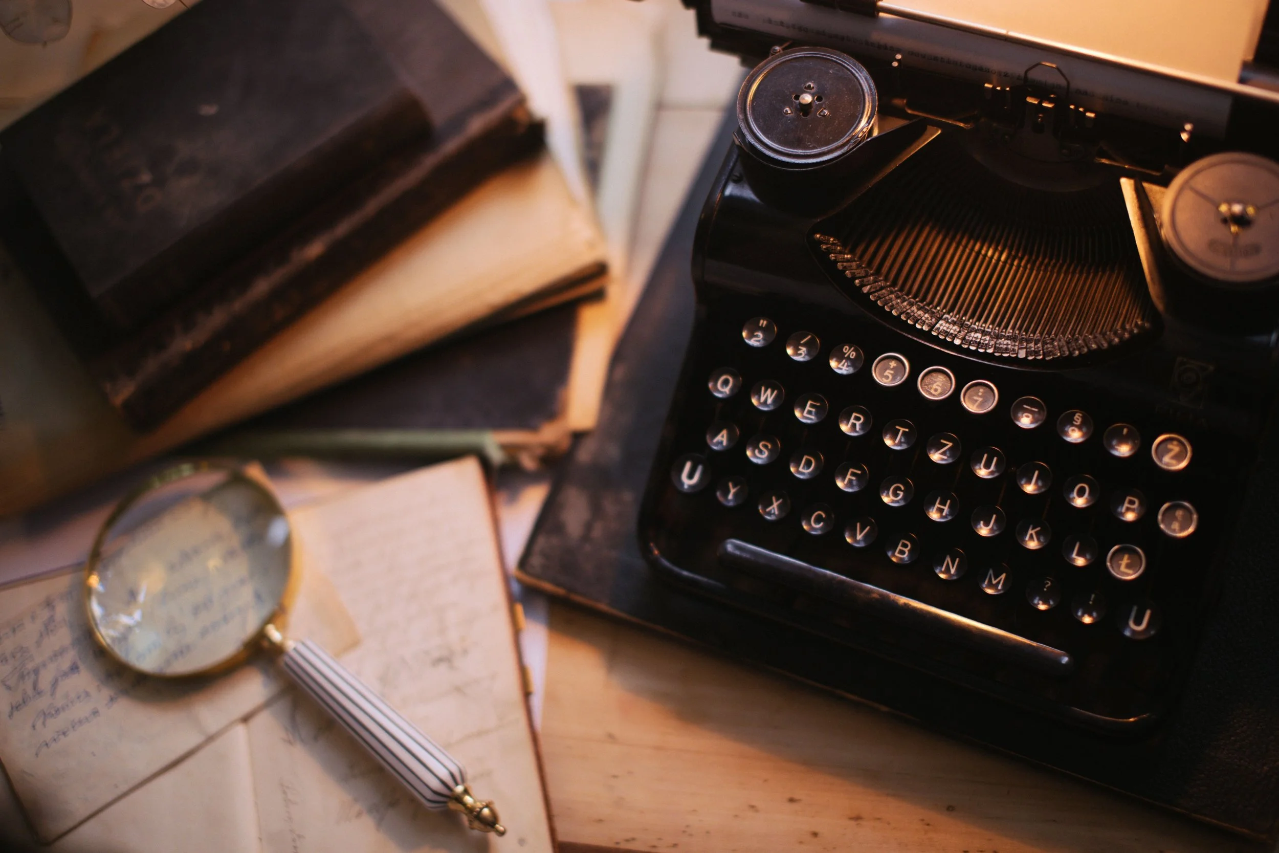 Close-up of a vintage typewriter, stack of old books, and a magnifying glass with handwritten notes on paper.