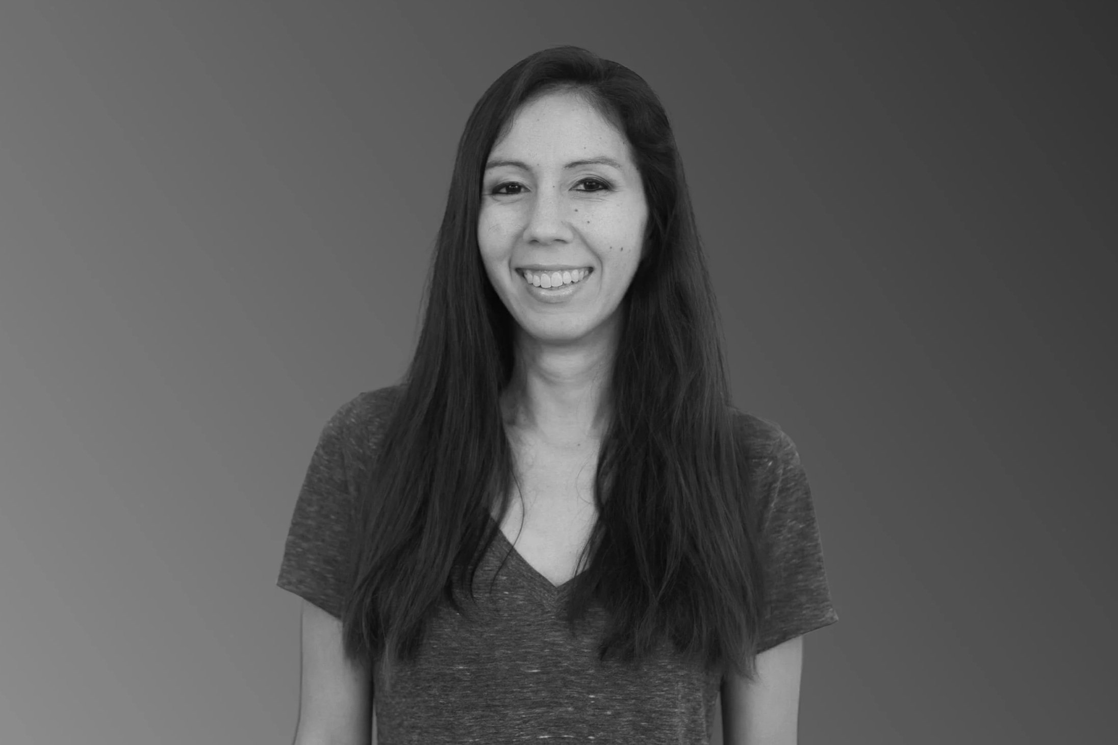 Black and white portrait of a woman with long dark hair, smiling, wearing a V-neck shirt, against a gradient background.