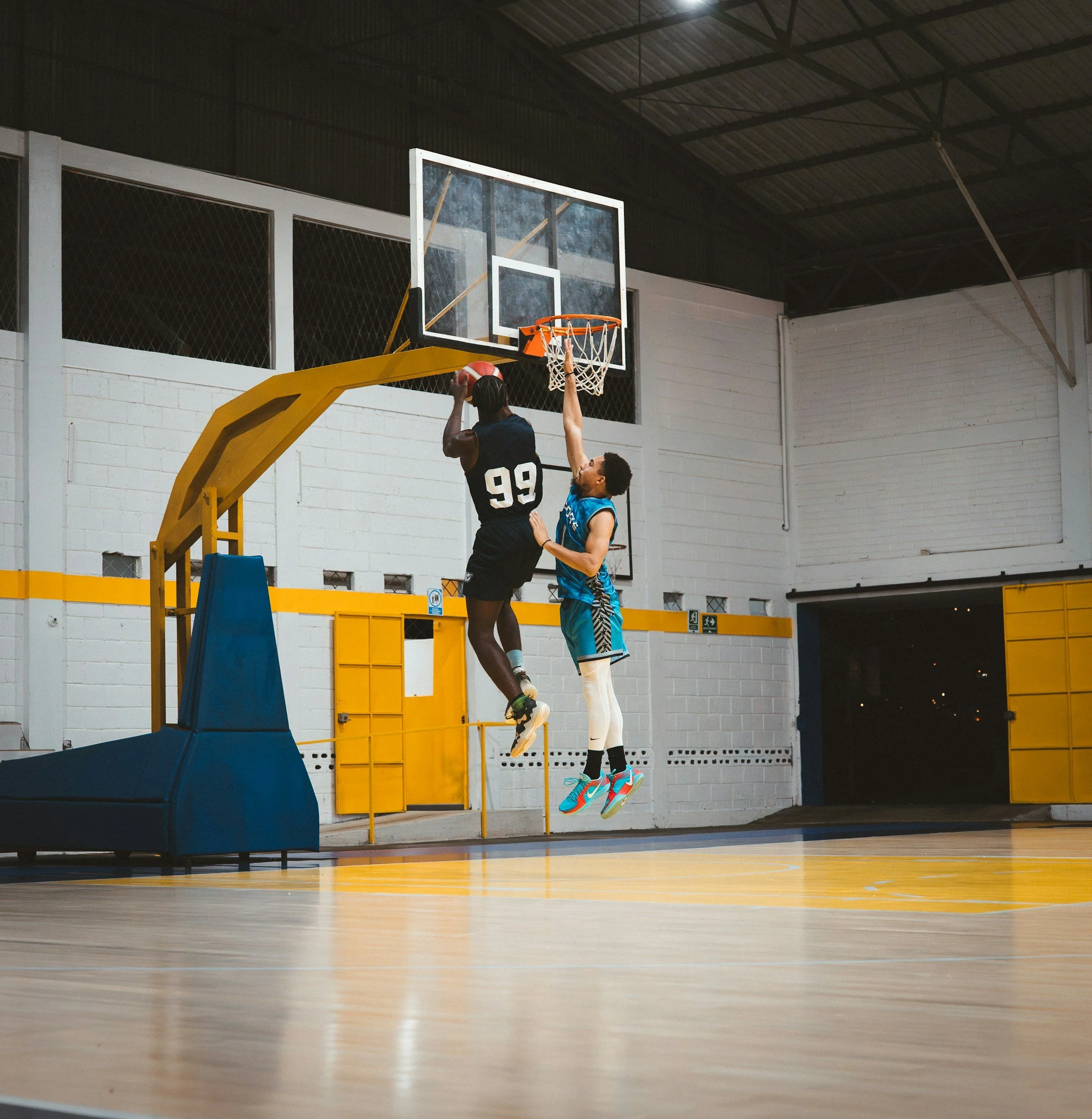 Two basketball players are jumping near the hoop, with one player in a black uniform attempting a shot and the other in a blue uniform defending.