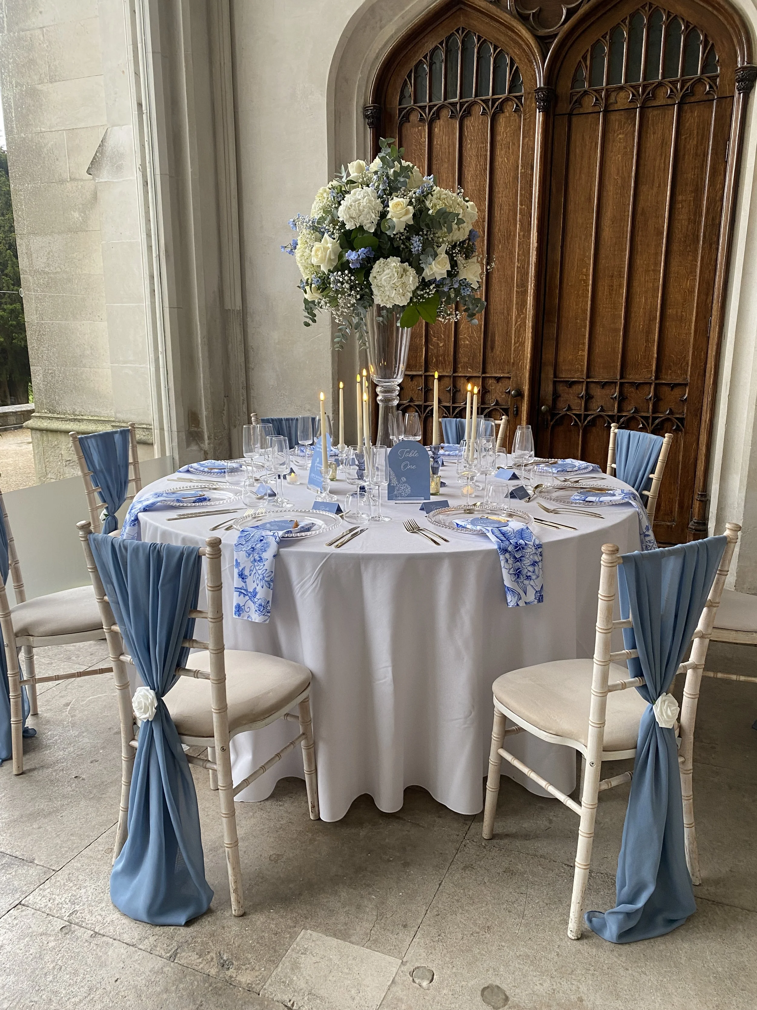 Wedding table with blue botanical napkins, blue chair drapes and a tall white and blue floral centrepiece.