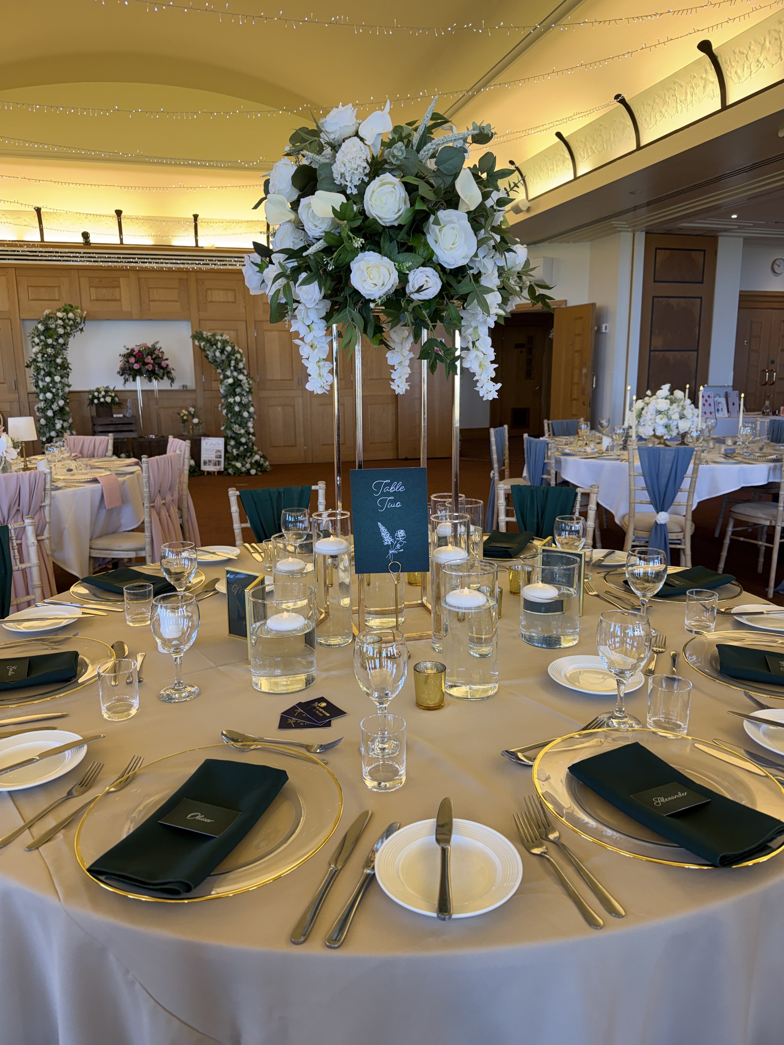 Round wedding table styled with forest green napkins and chair drapes, gold-rimmed chargers, floating candles and a tall white floral centrepiece.