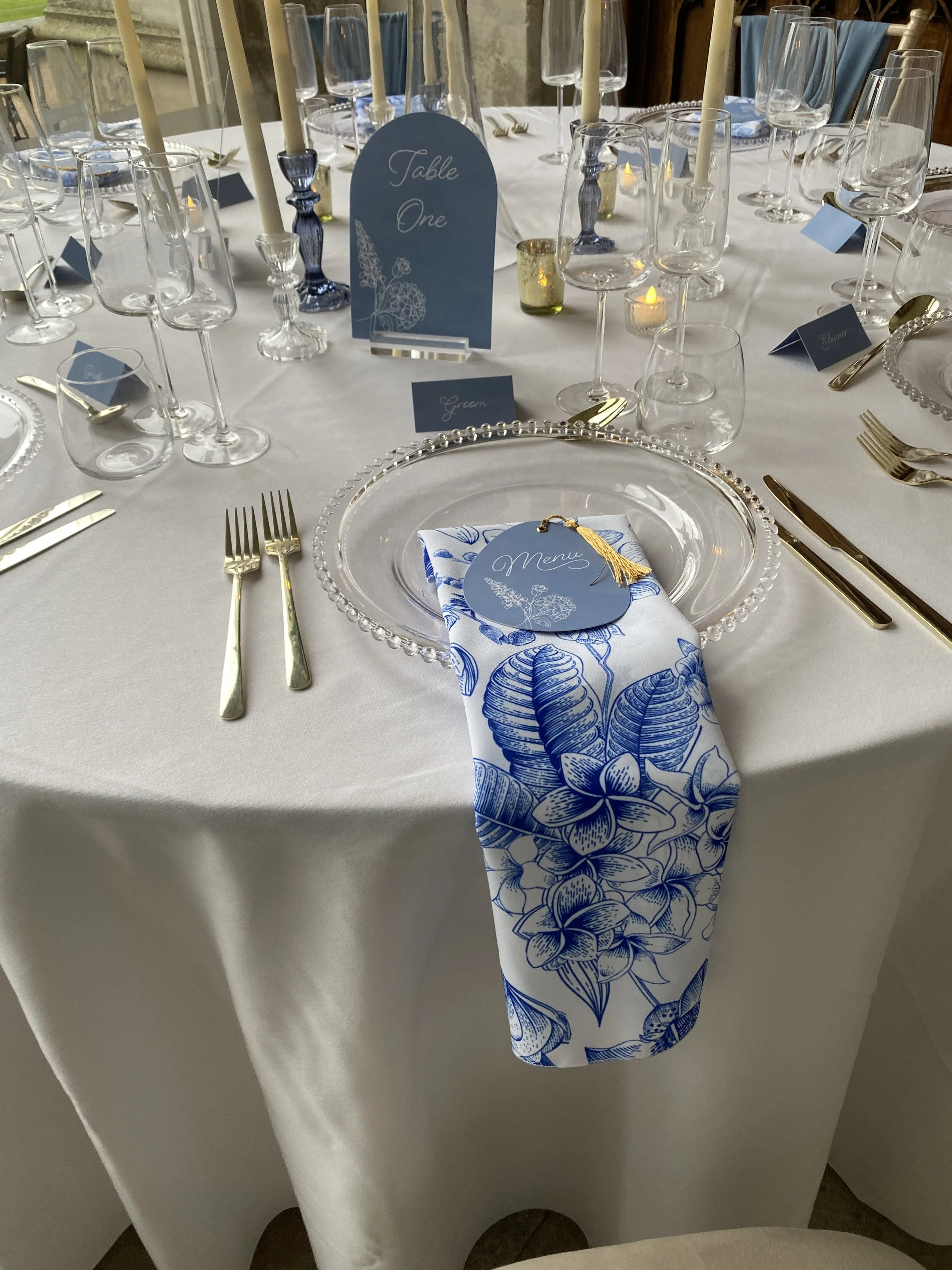 Close-up of a blue floral print polyester napkin beneath a blue menu with a gold tassel on a beaded glass charger.