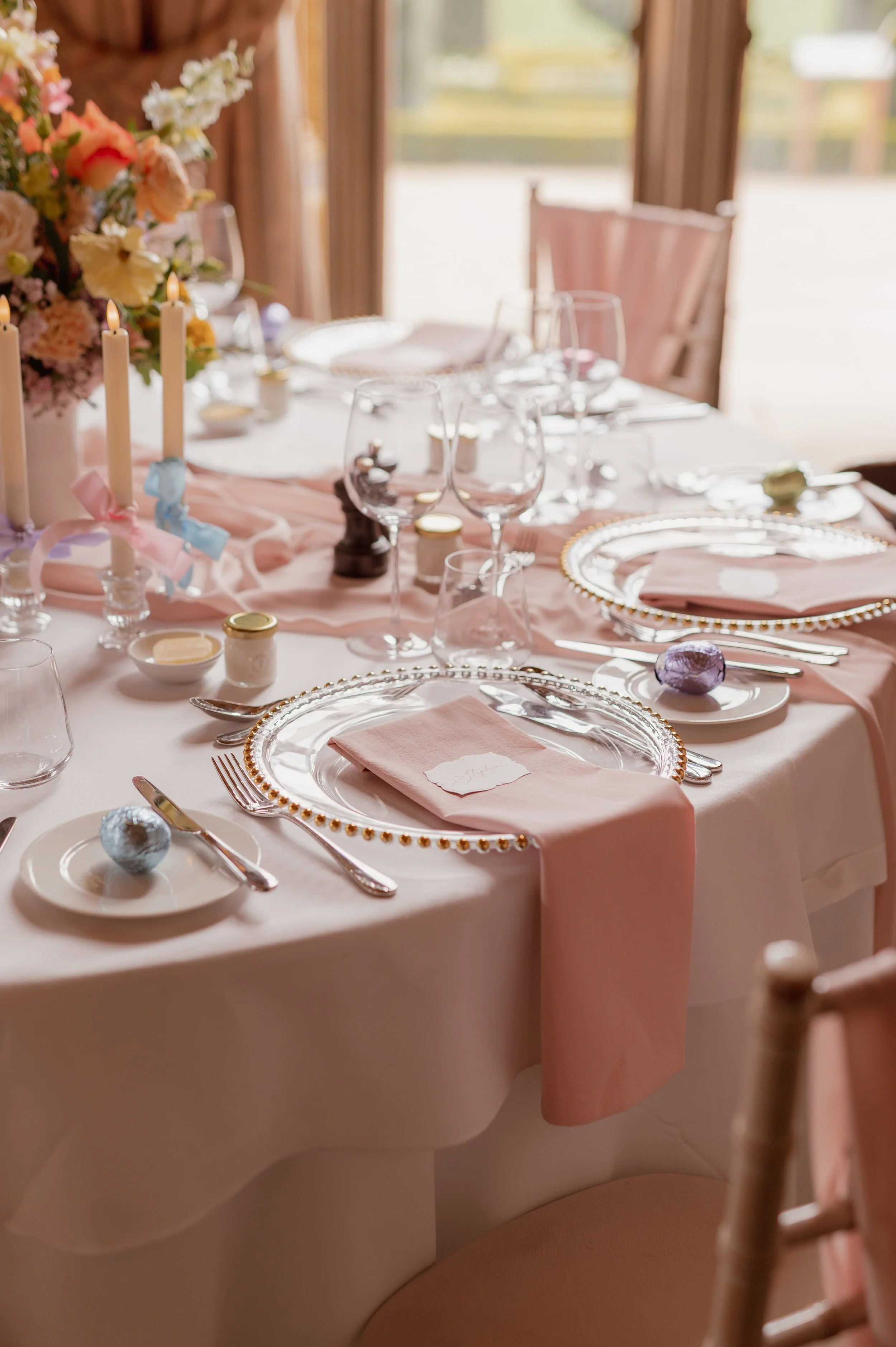 Pale pink cotton napkins styled with gold-beaded chargers, pastel flowers and ribbon-tied candles at Ashridge House.