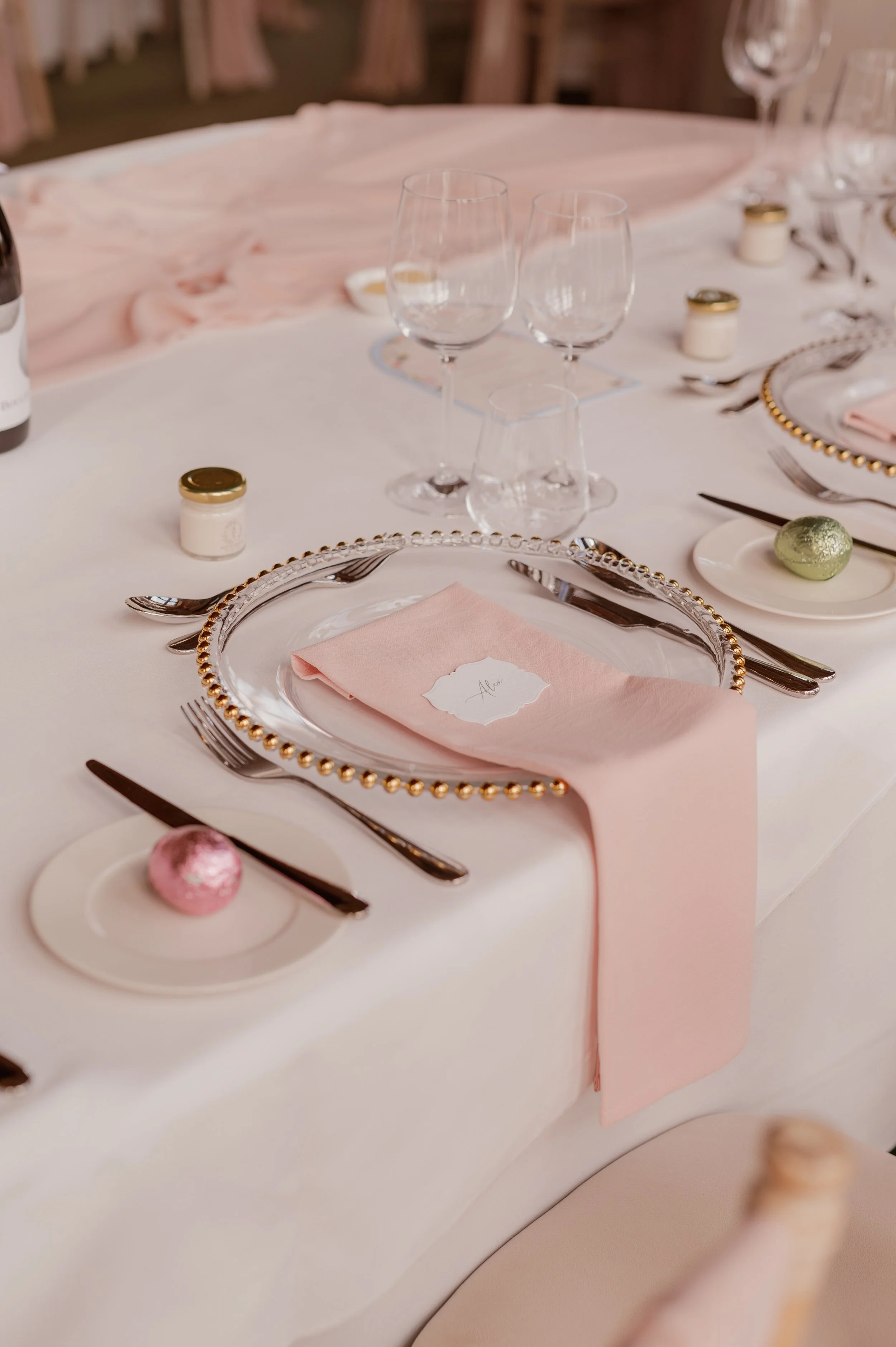Pale pink cotton napkin draped over a glass charger and table edge, with silver cutlery and clear wedding glassware.
