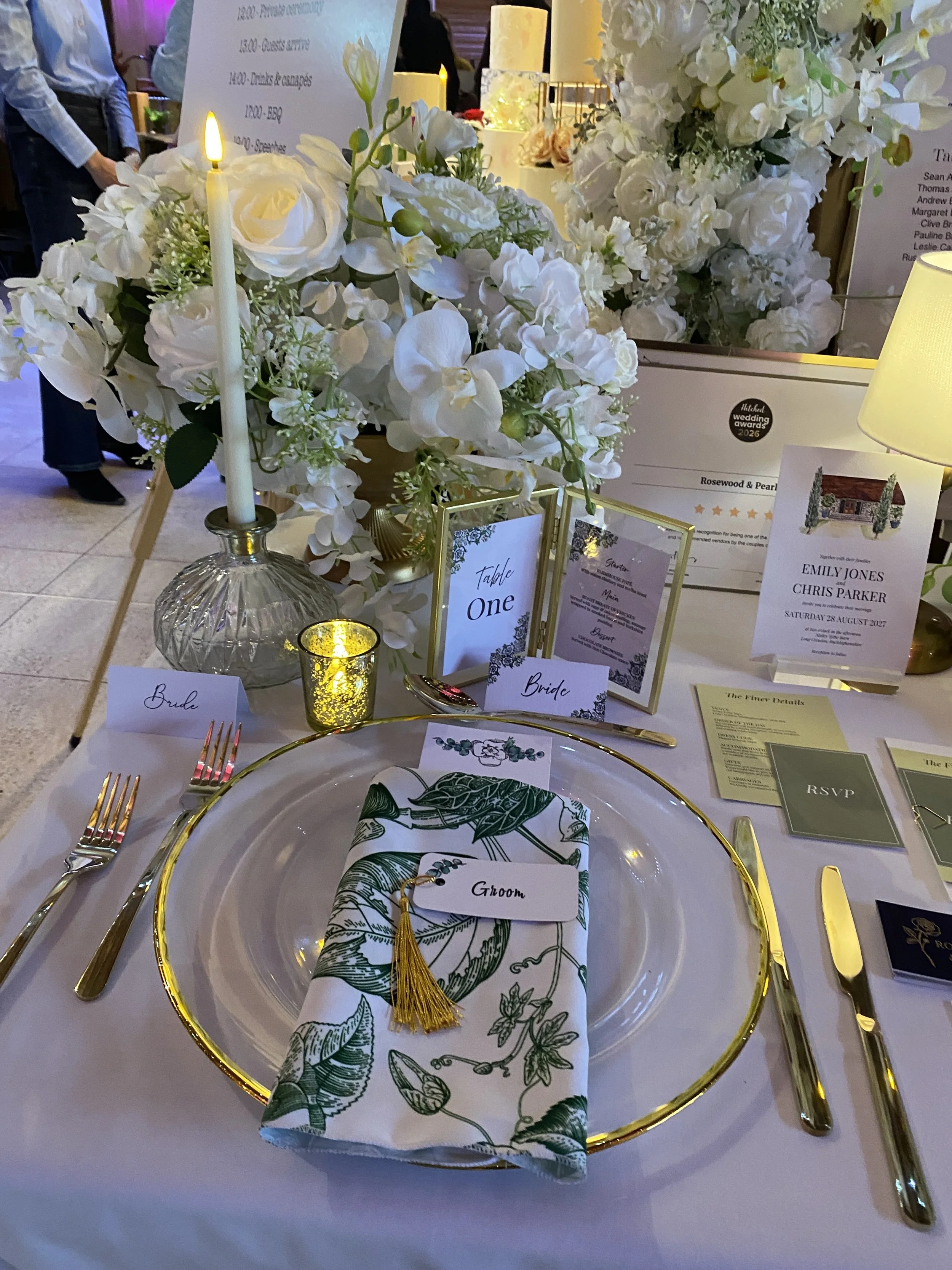 Green and white botanical napkin folded on a gold-rimmed glass charger, with gold cutlery, white roses and candlelight.