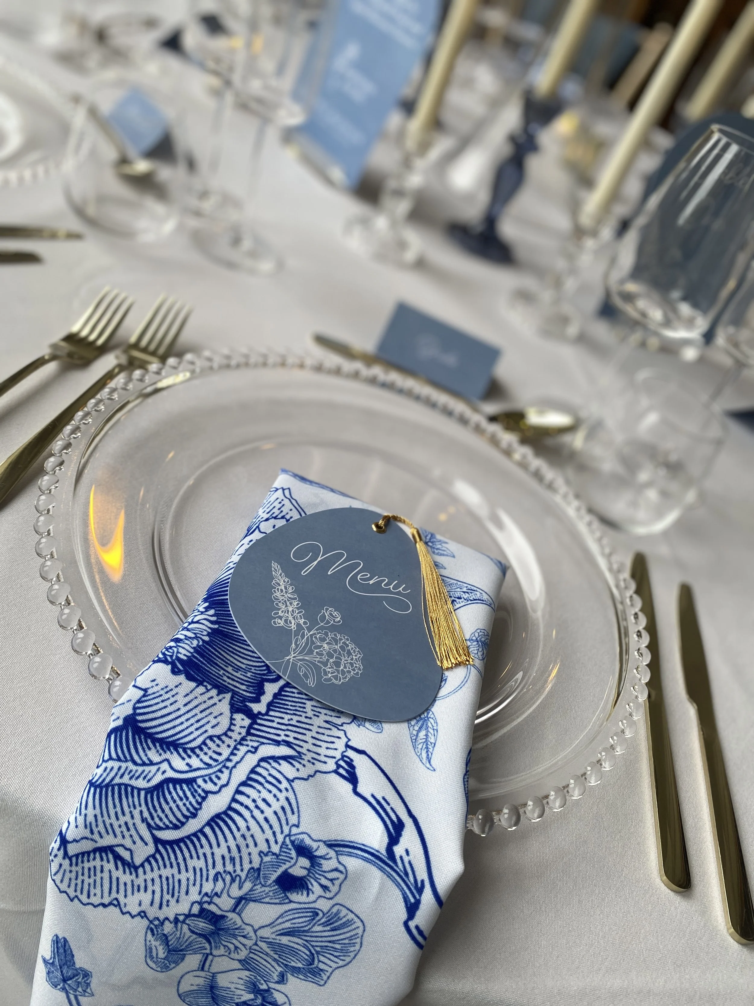 Blue and white botanical napkin draped over a glass charger, styled with a blue menu and gold cutlery.