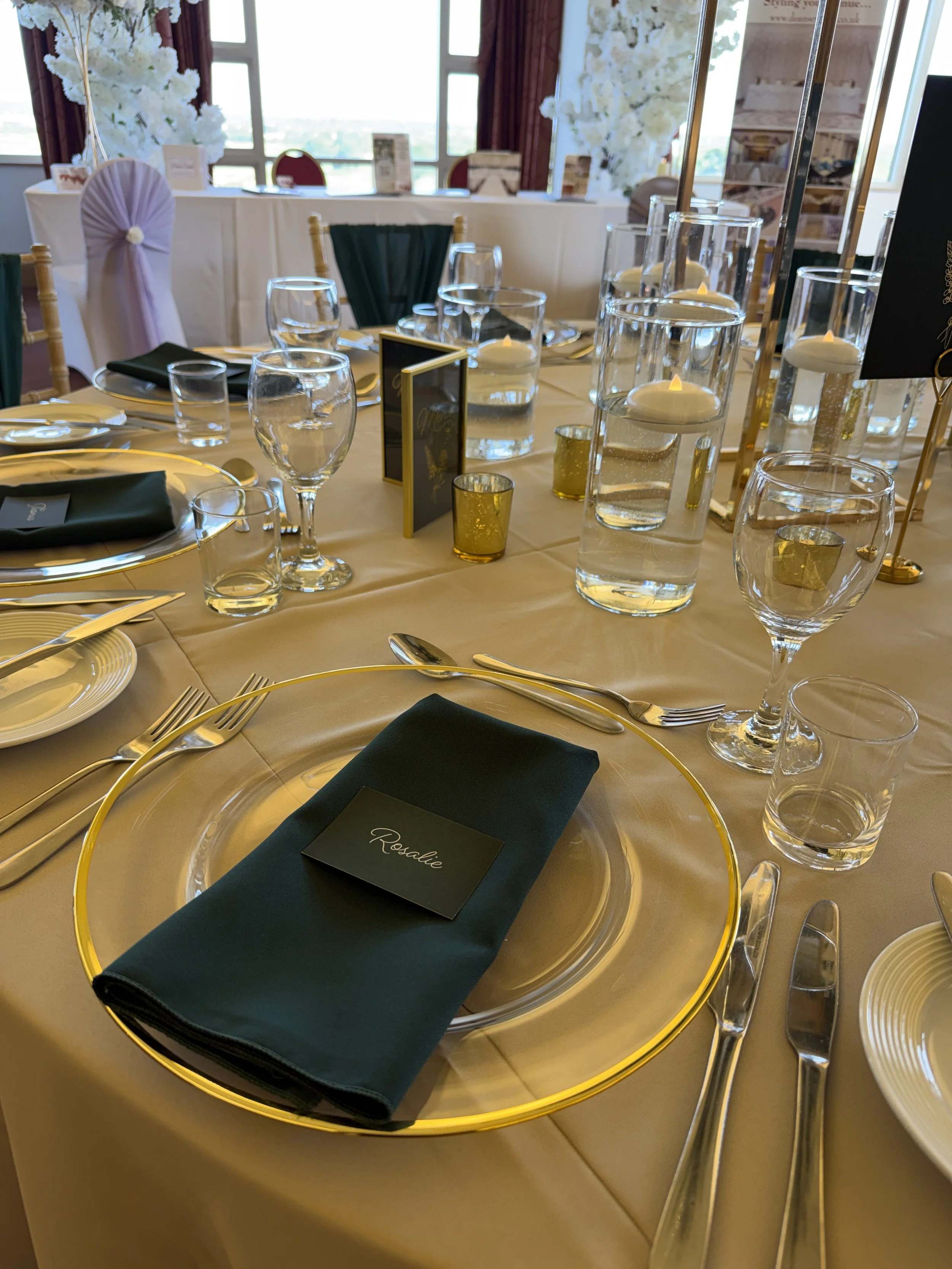 Forest green polyester napkin folded on a gold-rimmed glass charger with a matching place card, silver cutlery and clear glassware.