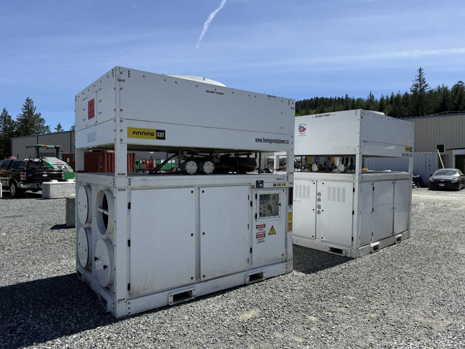 Three large white industrial cooling units on a gravel lot, with a background of parked cars, a building, and green hills under a clear blue sky.