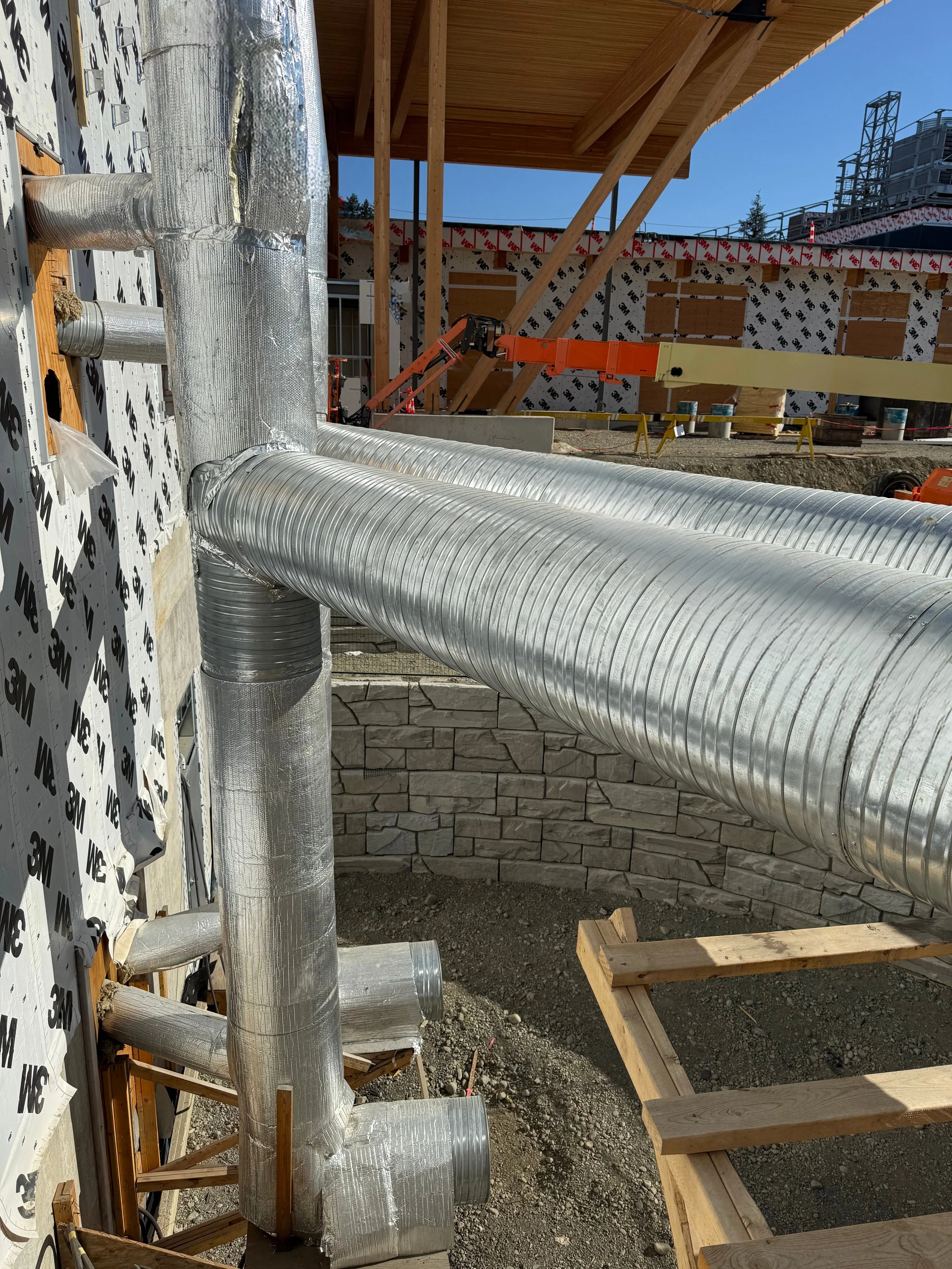 Construction site with large silver HVAC ducts, wooden framing, and partially built stone wall under a sunny sky.