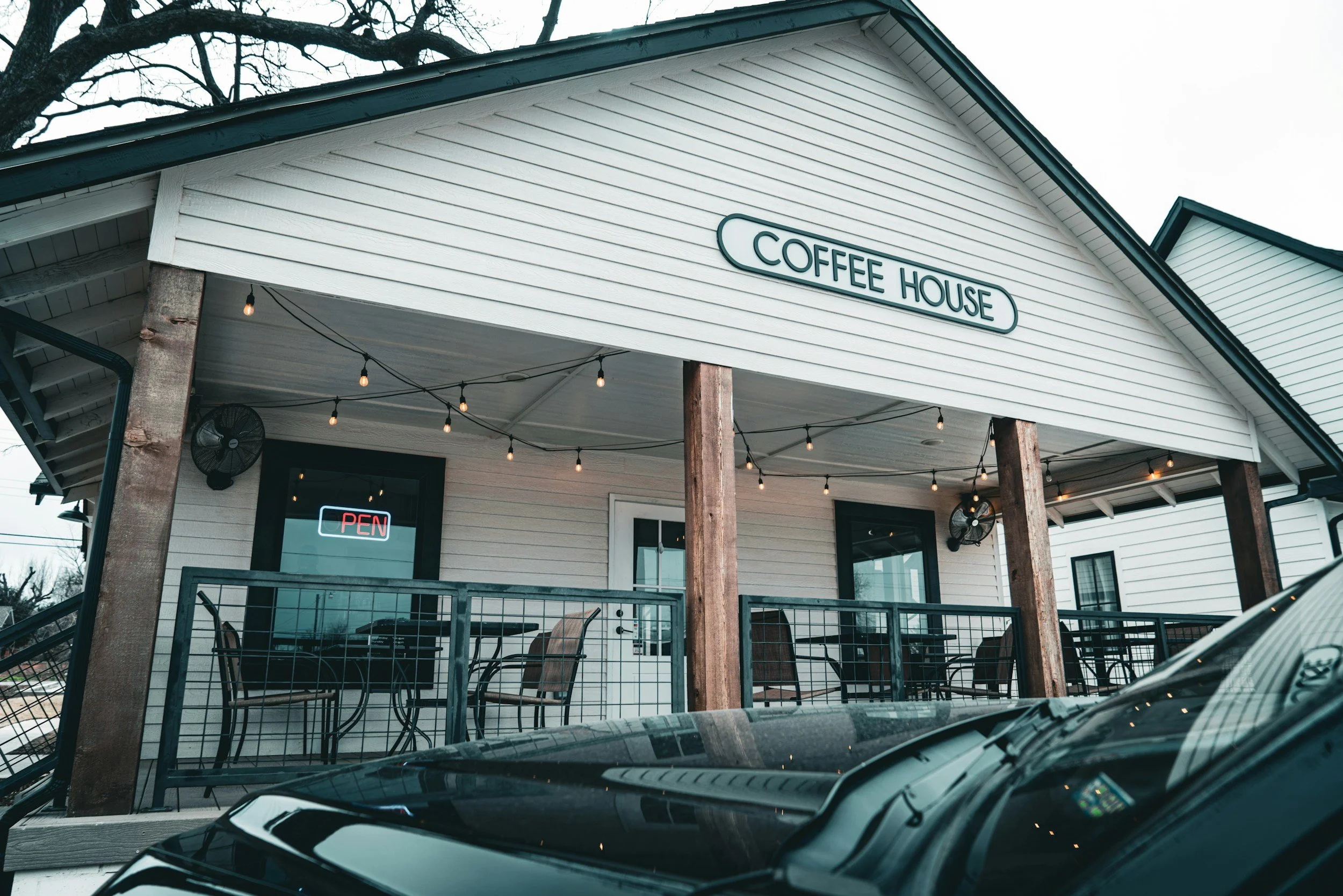Exterior view of a coffee house with a white exterior, black-framed windows, and digital sign that says 'OPEN'. There is outdoor seating with tables and chairs, string lights hanging from the porch ceiling, and a car parked in front.