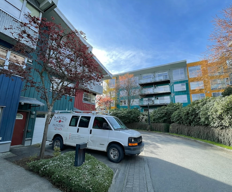 A parking area with a white van labeled 'Cascade Painting' parked in front of colorful apartment buildings with red, blue, green, yellow, and orange facades, surrounded by trees and shrubs under a partly cloudy sky.