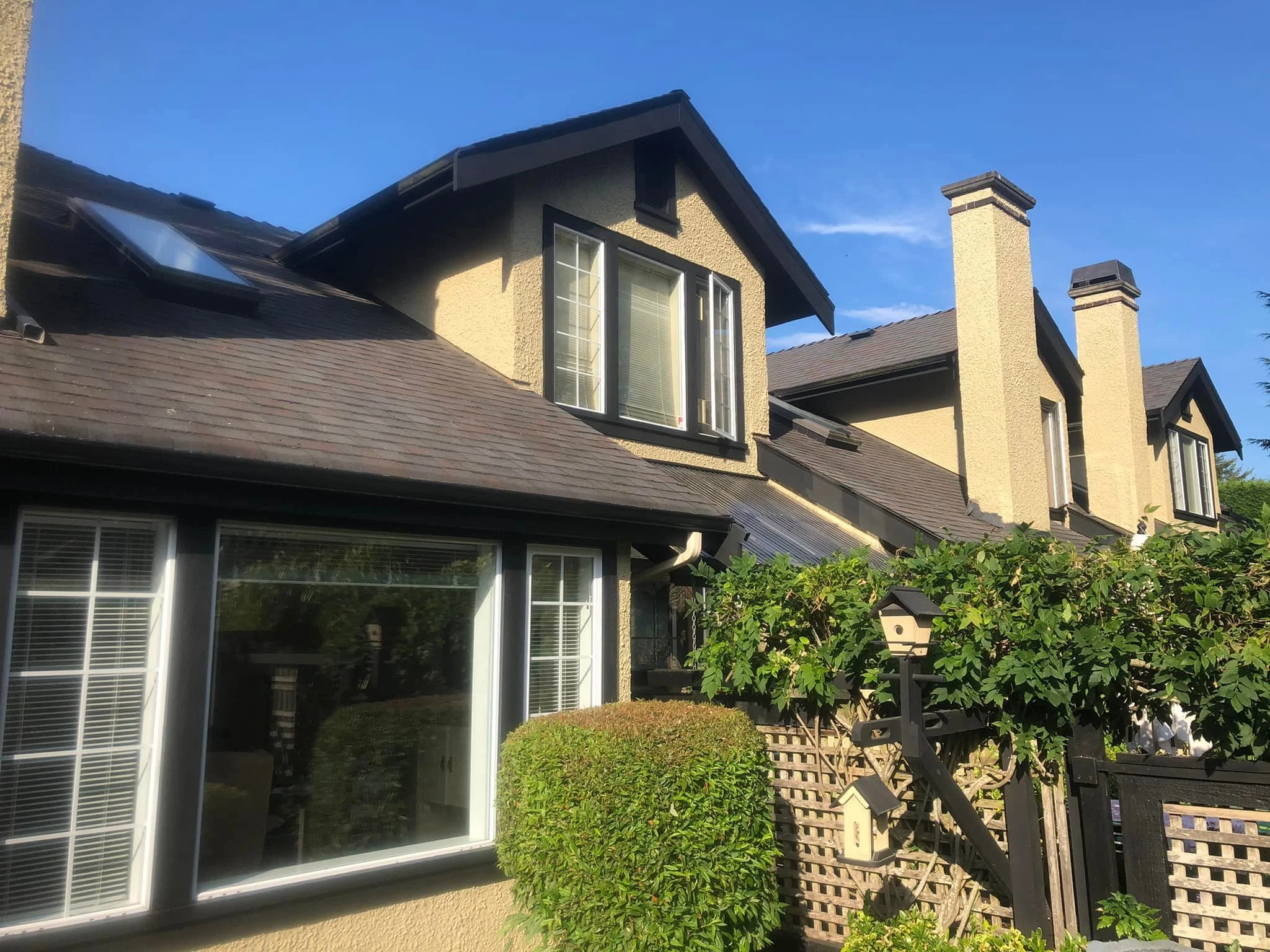 A multi-story house with beige stucco walls, a dark gray shingle roof, and multiple chimneys. There are large windows on the upper and lower levels, some with blinds. A lush green hedge and wooden lattice fence with birdhouses are in the foreground. The sky is clear and blue.