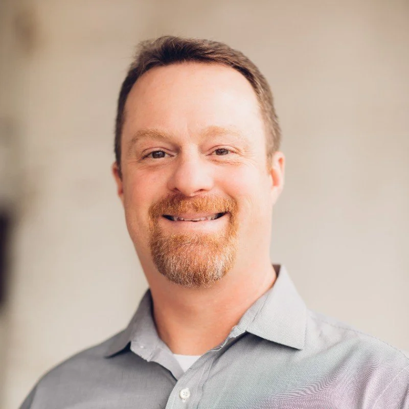 A smiling man with a goatee and short brown hair, wearing a gray collared shirt, posing in front of a neutral background.