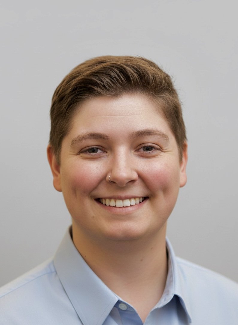 Close-up portrait of a young person with short brown hair, smiling, wearing a light blue collared shirt against a plain light gray background.
