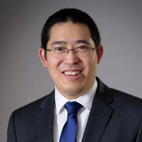 Professional headshot of a man wearing glasses, a dark suit, white shirt, and blue tie, smiling against a gray background.