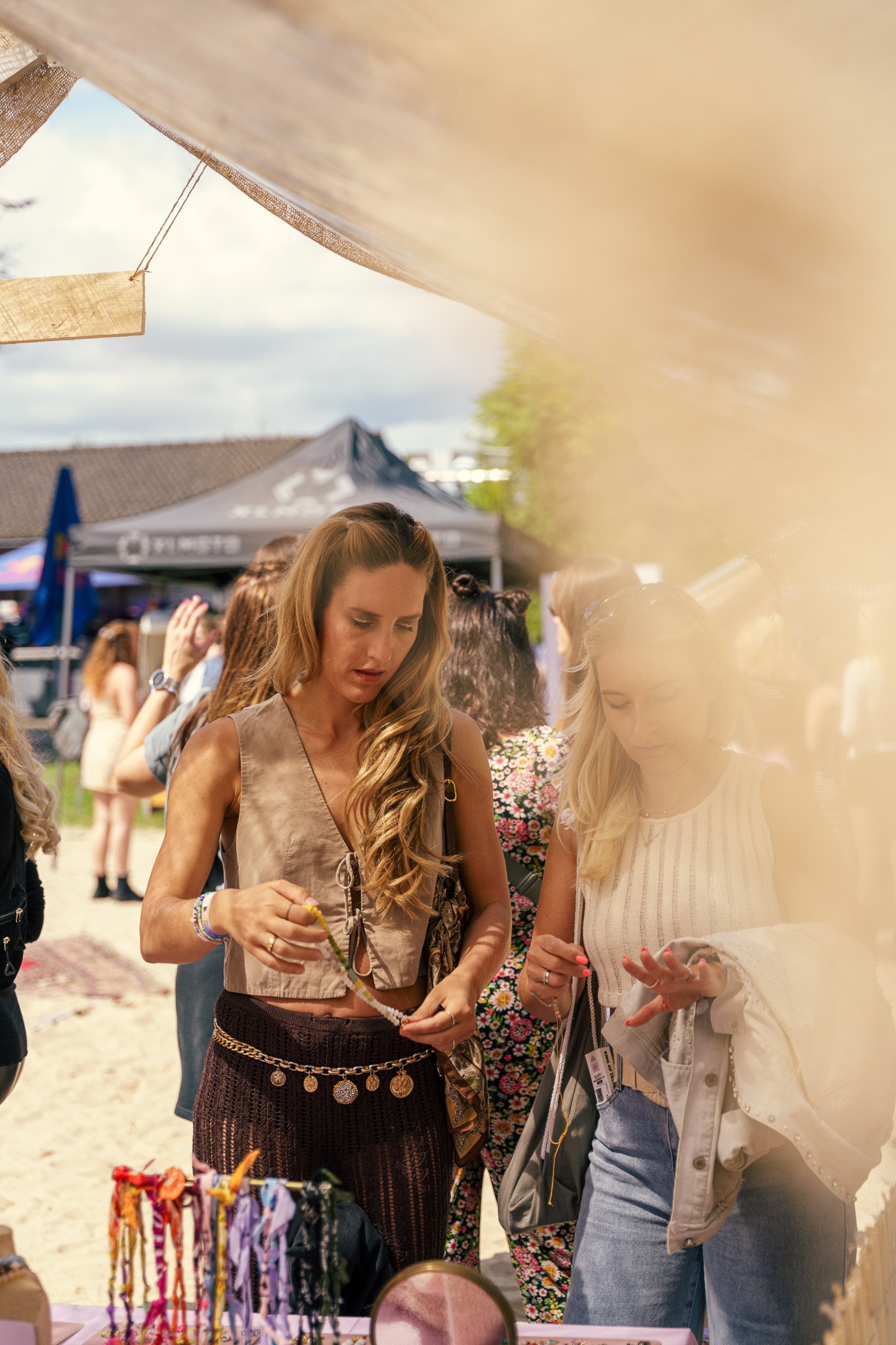 Two women shopping at an outdoor craft fair, browsing jewelry, with tents and other people in the background.