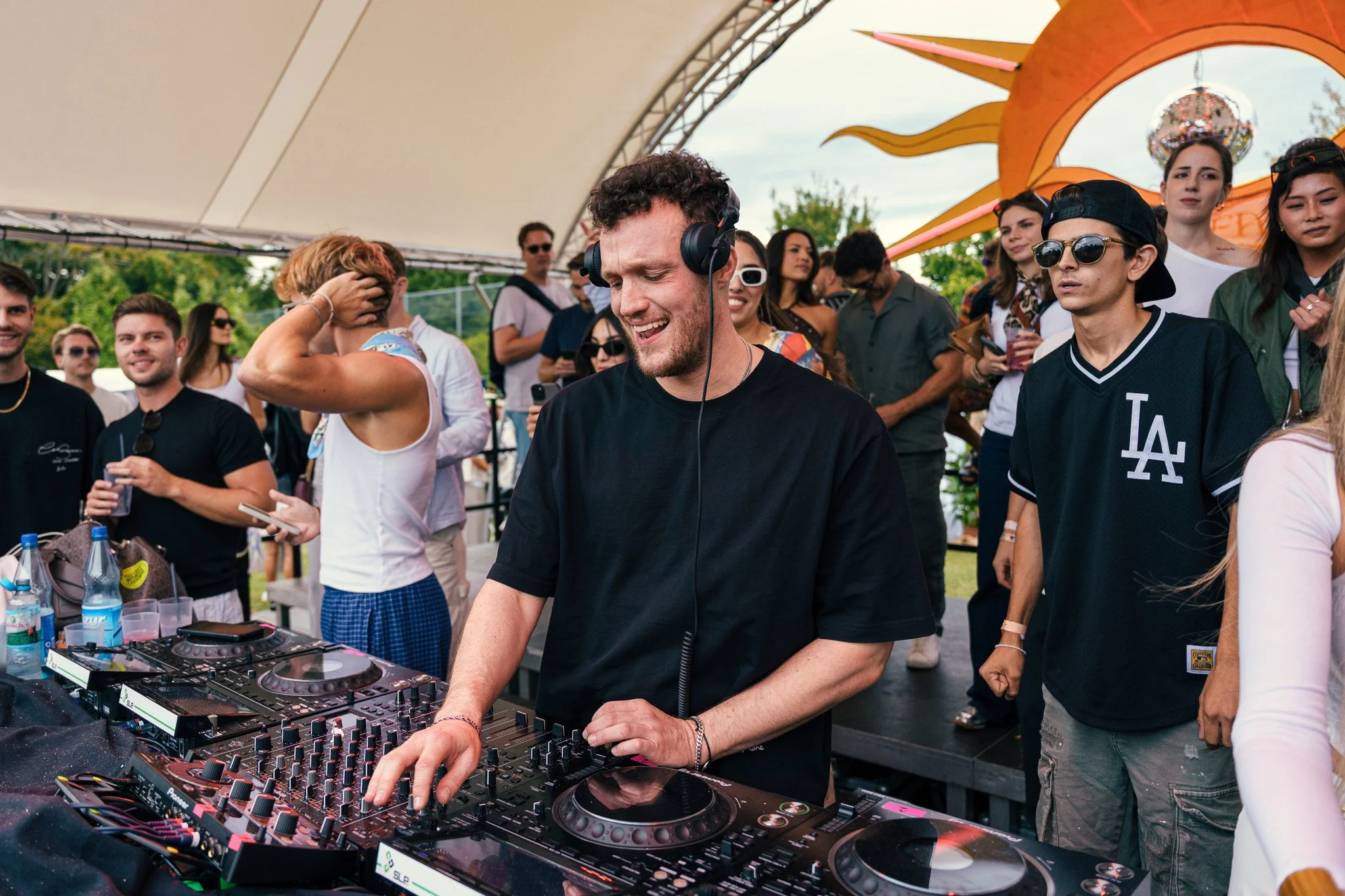 A male DJ with curly hair, wearing headphones and a black t-shirt, is mixing music with a DJ controller at an outdoor event. He is smiling and surrounded by a diverse crowd of people enjoying the music under a tent with a decorative orange and yellow
