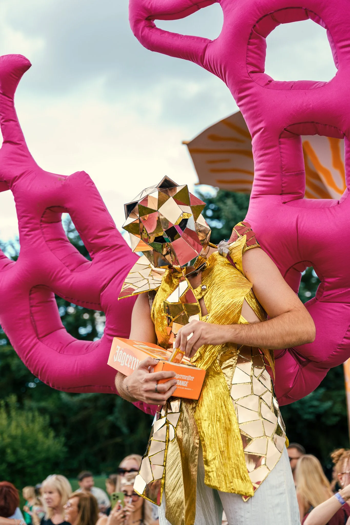 A person dressed in a gold metallic outfit with a geometric, reflective mask covering their face, holding an orange box labeled 'Jägermeister' during an outdoor event with a crowd in the background.