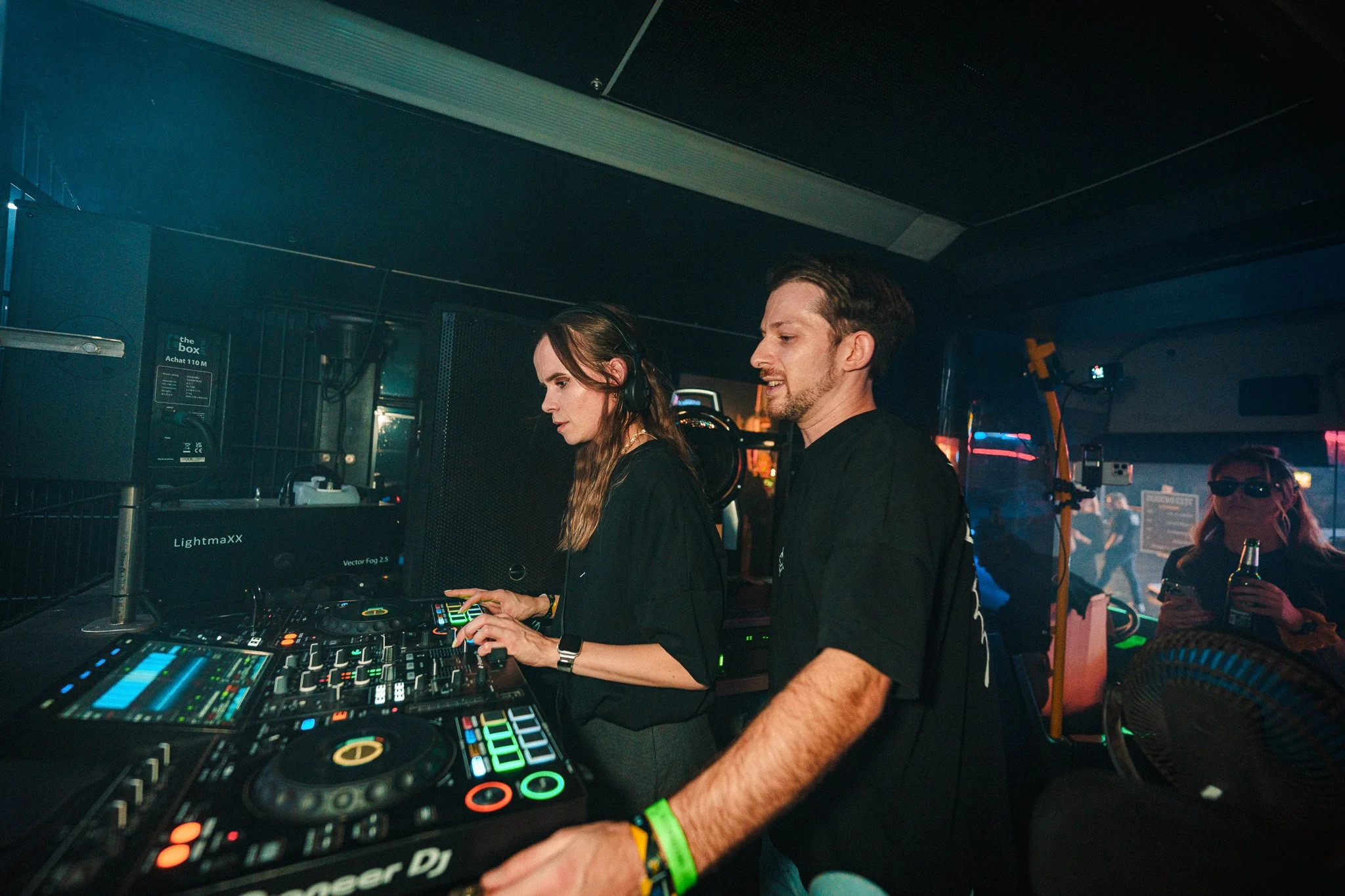Two DJs, a woman and a man, are inside a nightclub or music venue with DJ equipment, lighting, and people in the background, including a woman drinking from a bottle.