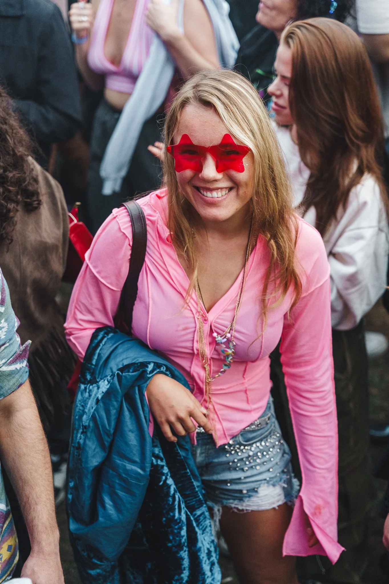 Smiling woman wearing red star-shaped sunglasses, pink hoodie, and denim shorts at a crowded outdoor event.