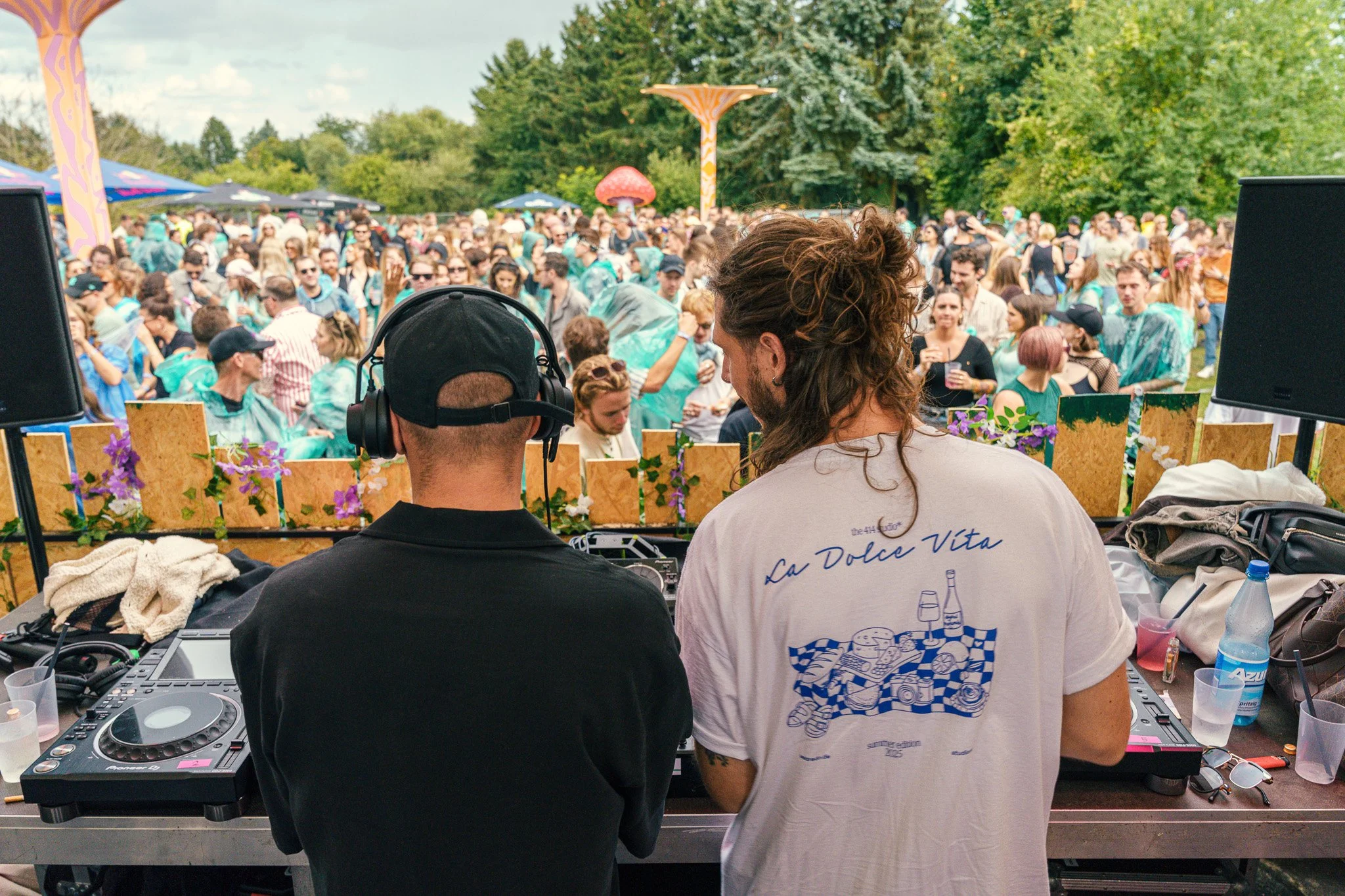 Two DJs perform at an outdoor music festival with a large crowd dancing in a green, wooded area.