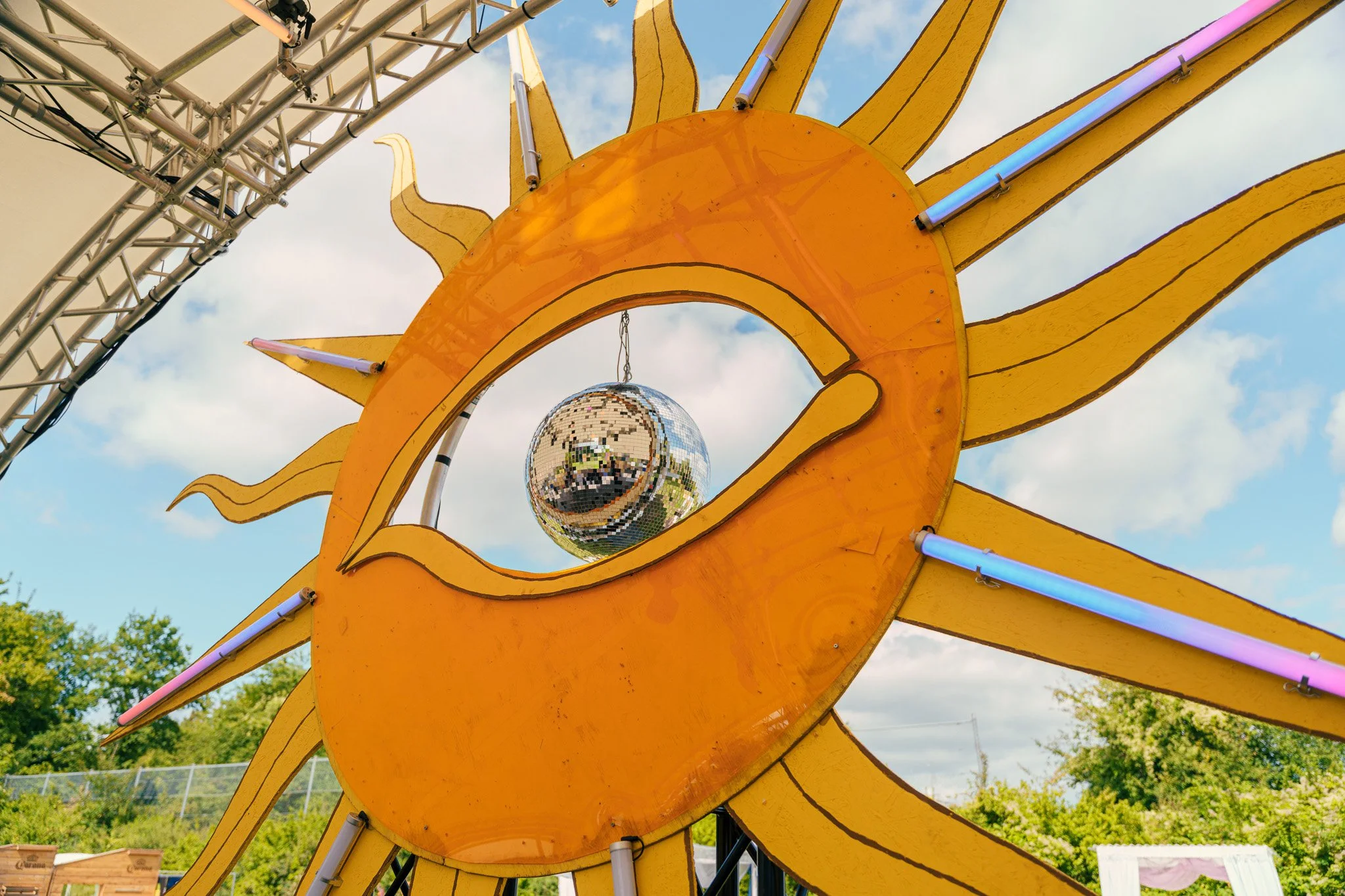 Yellow sun-shaped carnival ride with a reflective disco ball hanging in the center, set outdoors with trees and a blue sky in the background.