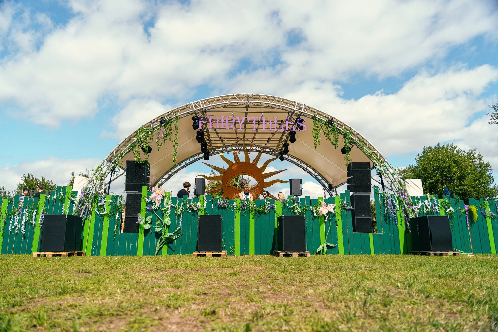 Outdoor music festival stage decorated with green and purple flowers, with a large sun and eye emblem at the center, under a partly cloudy sky.