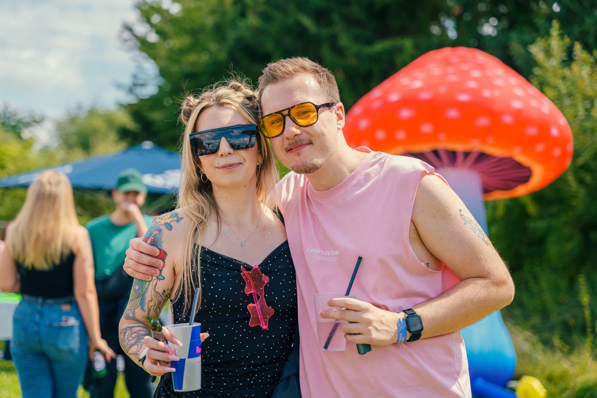 A young man and woman are posing outdoors at a festival or fair, each wearing sunglasses and holding drinks. In the background, there are green trees, blue and orange umbrellas, and a large red and white mushroom-shaped decoration.