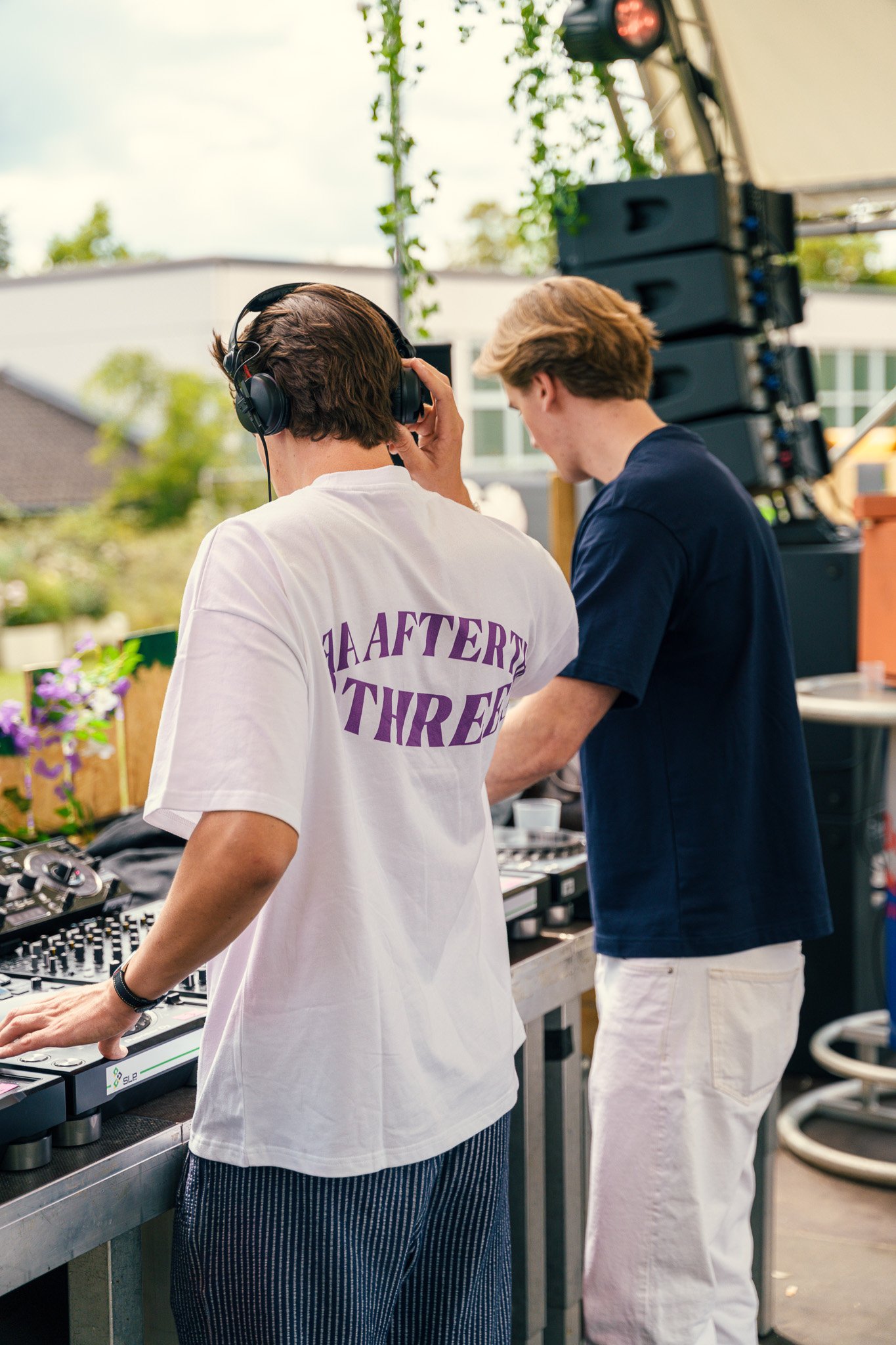 Two young men DJing at an outdoor event with DJ equipment and large speakers, one wearing headphones and a white T-shirt, the other in a navy T-shirt and white pants.