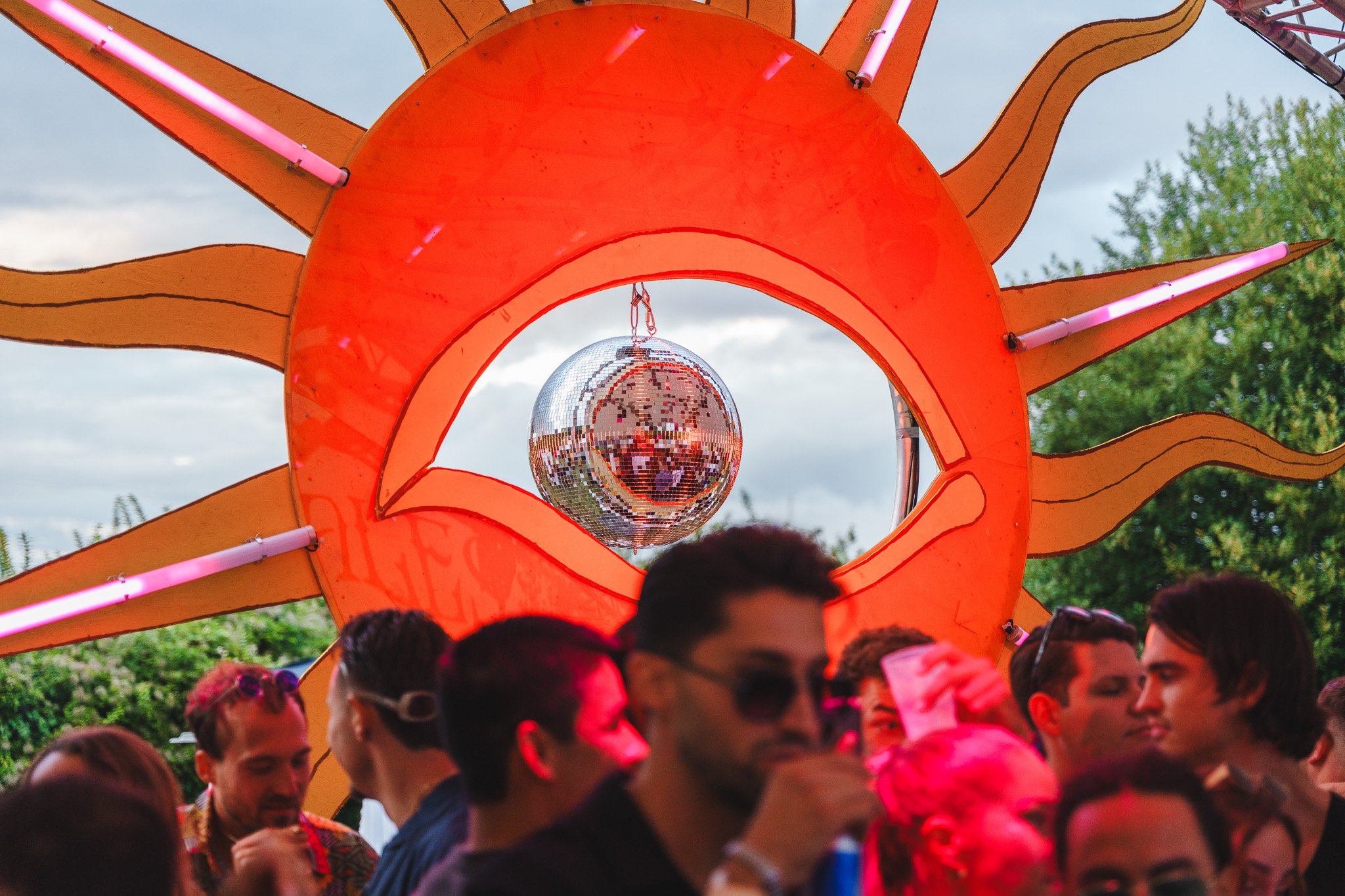 People at a party or festival under a large decorative sun with a mirror disco ball hanging in the center.