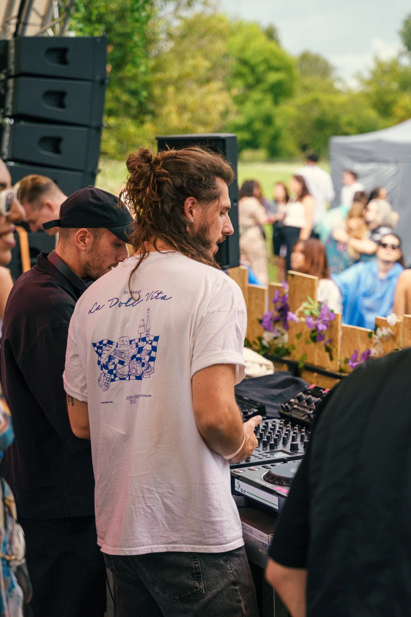DJ with long hair playing music at an outdoor event, surrounded by people and greenery.