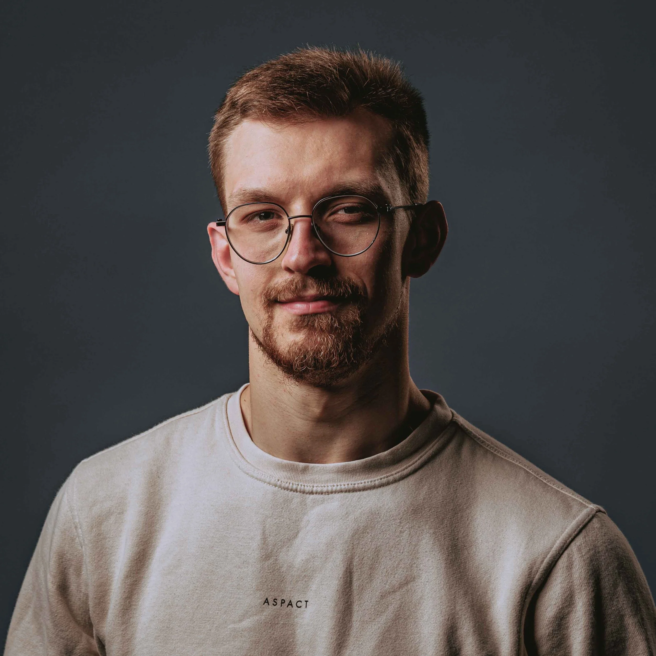 Portrait of a young man with short brown hair, wearing round glasses, a goatee, and a beige T-shirt with the word 'ASPACT' on it, against a dark gray background.