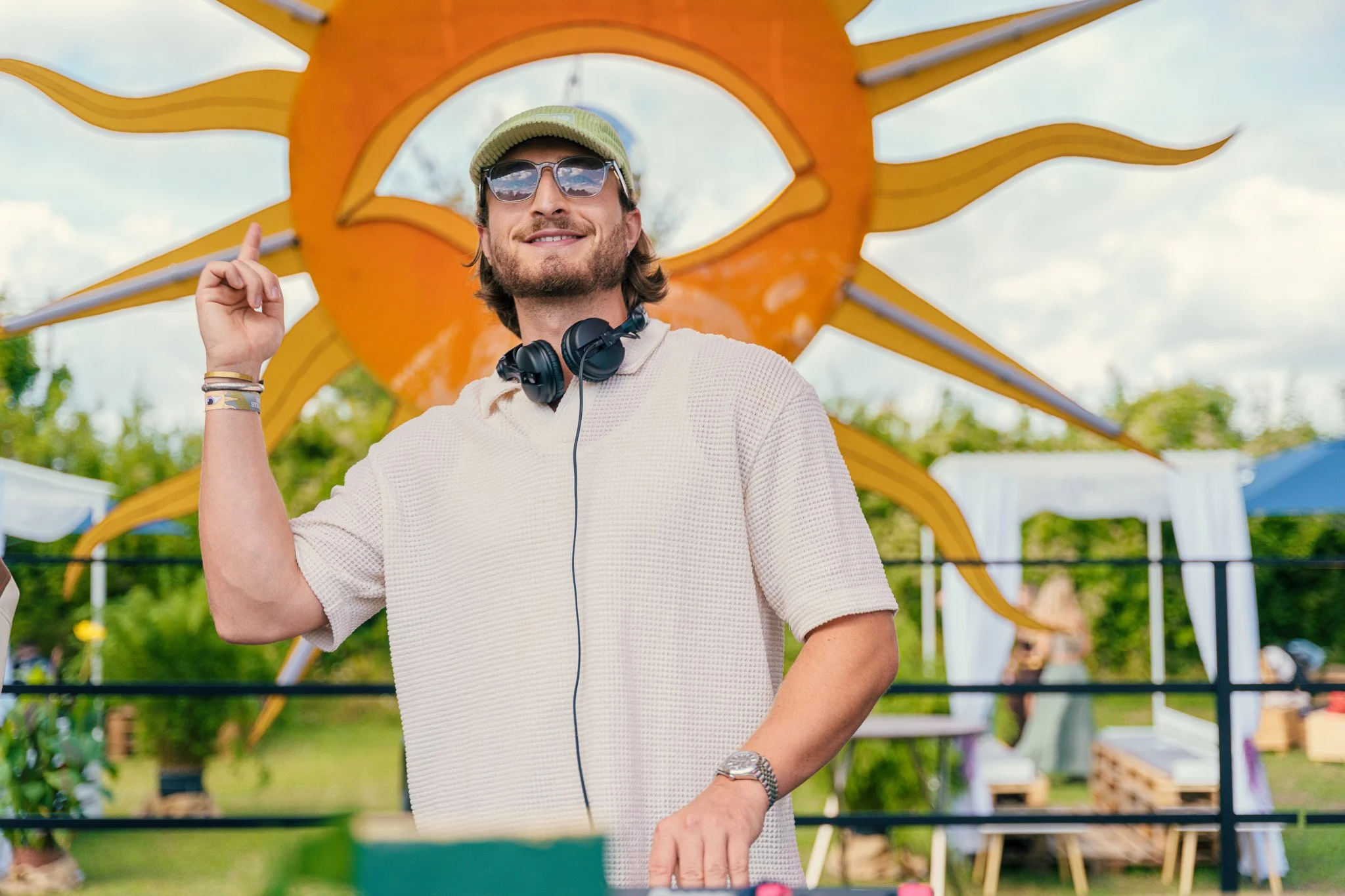 A man with long hair, wearing sunglasses, a light-colored knit shirt, a watch, and headphones around his neck, is smiling and pointing upwards with his right index finger. Behind him is a large orange and yellow sun-shaped decoration at an outdoor ev