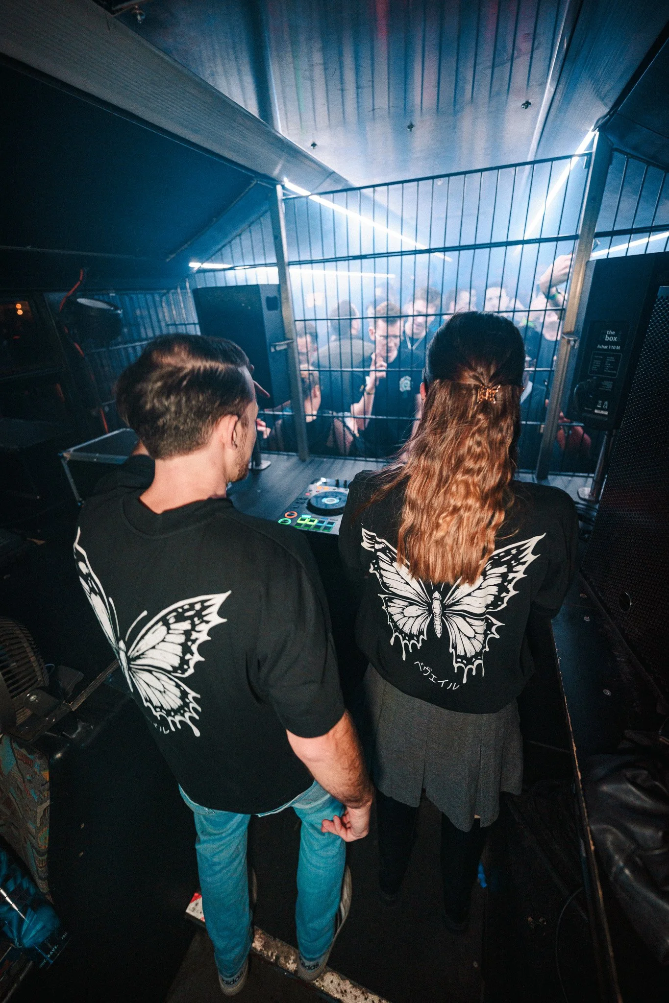 Two DJs, a man and a woman, stand behind a DJ mixer at a nightclub, facing a crowd of people dancing behind a metal barrier. Both wear matching black shirts with large butterfly designs on the back.