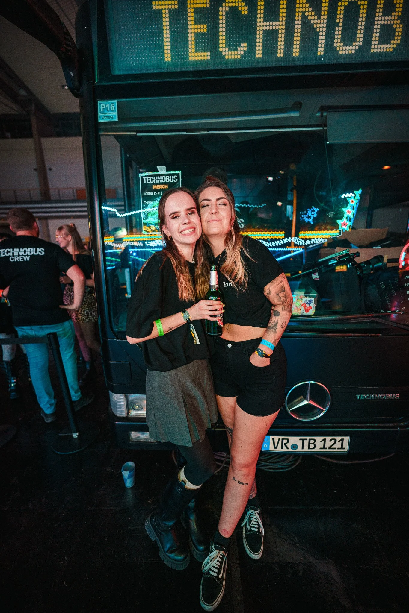 Two women smiling and posing together at a music event or party, one holding a beer, standing in front of a black Mercedes-Benz vehicle with bright neon lights and sign reads 'TECHNO'.