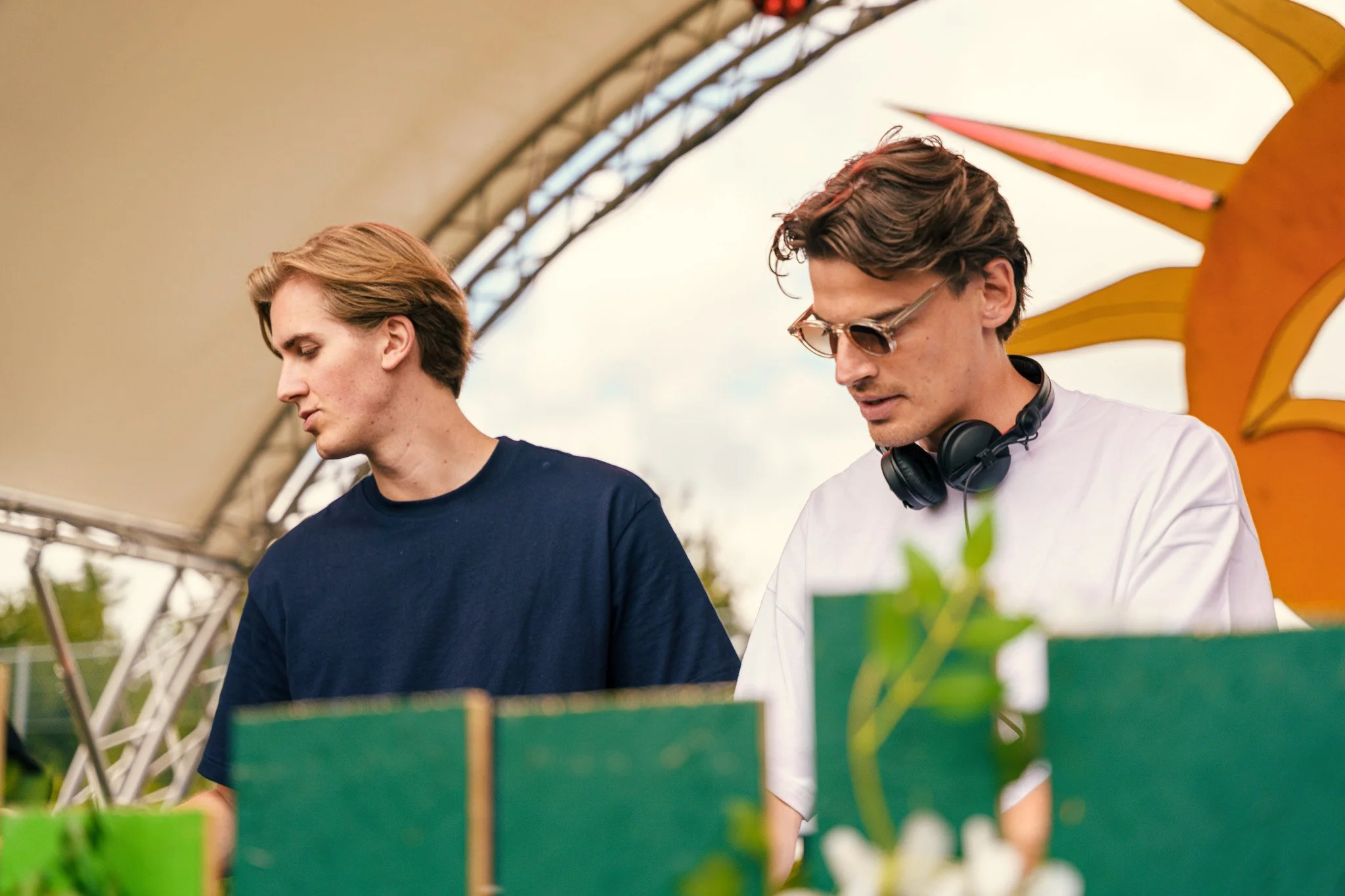 Two young men at an outdoor event or market under a canopy, looking down at a table with green and other colored boxes. The man on the right wears sunglasses, a white T-shirt, and headphones around his neck, while the man on the left wears a navy blu