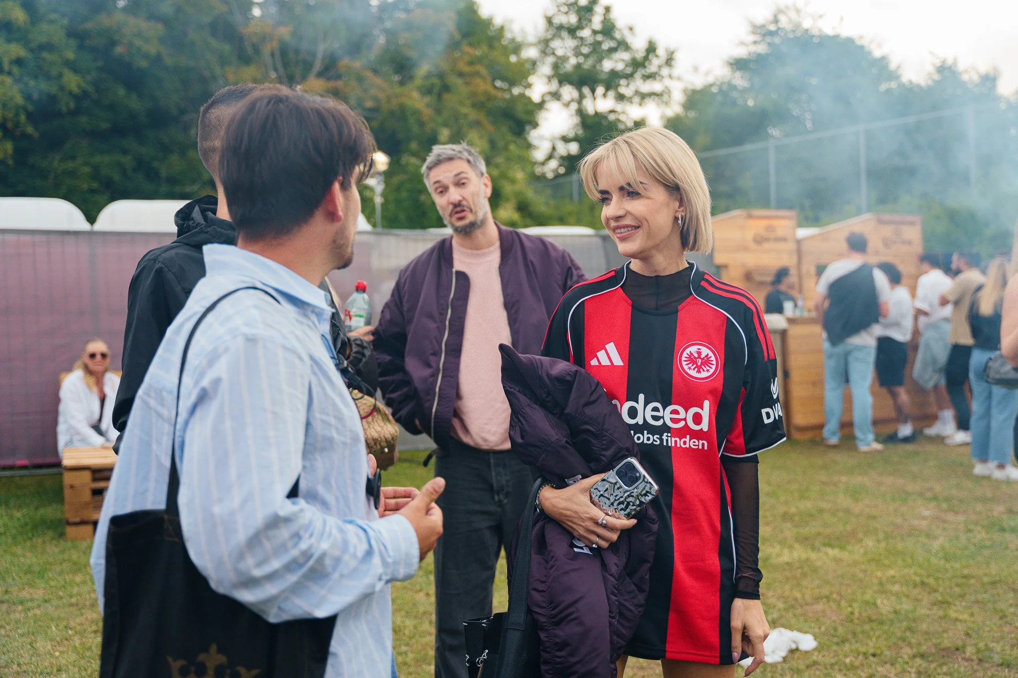 A woman wearing a black and red soccer jersey with an emblem, smiling and holding a phone and jacket, engaged in conversation with a young man in a blue striped shirt at an outdoor event. Others are in the background with green trees and a crowded se